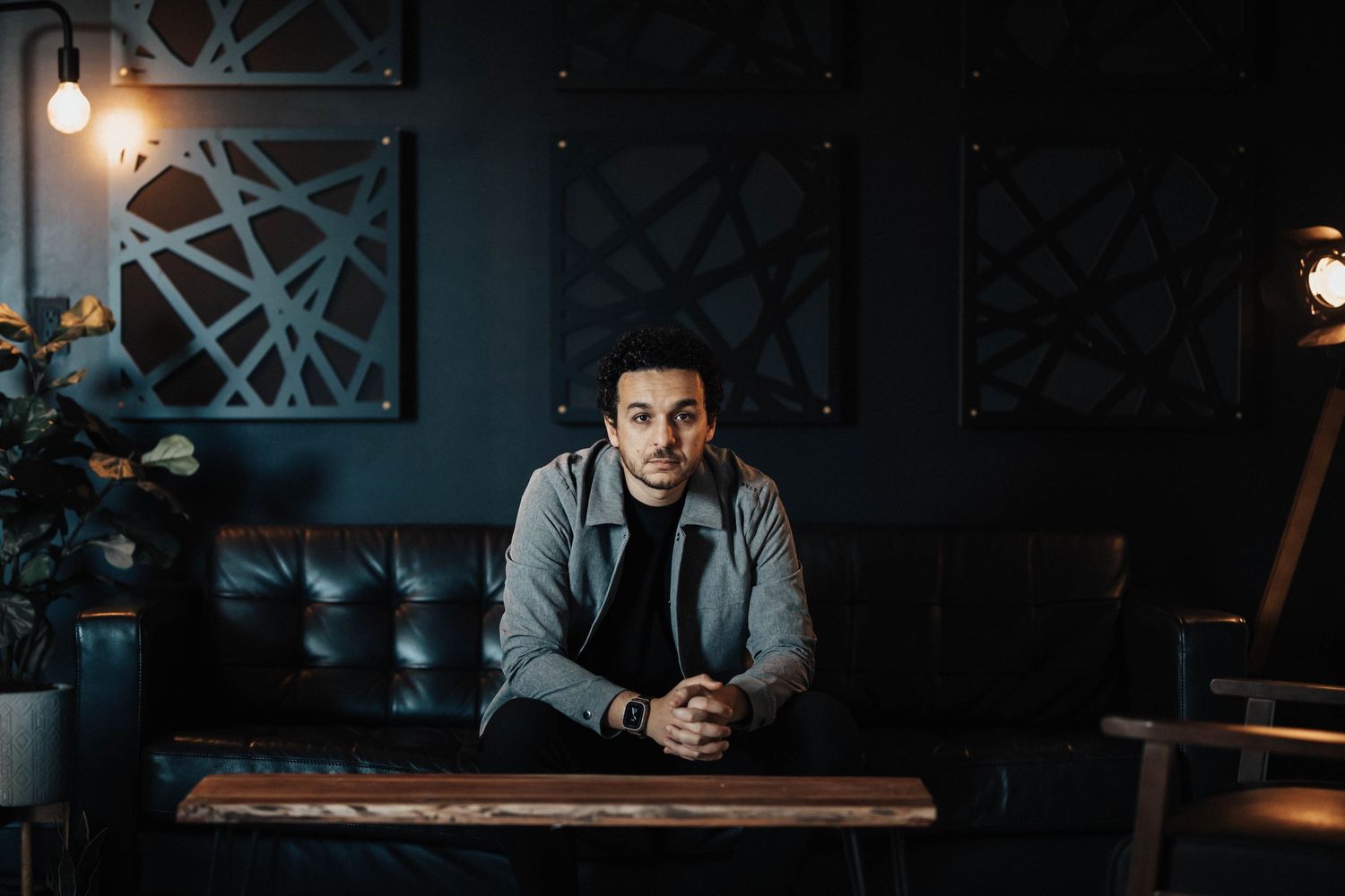 Young man sitting on leather sofa in moody dark interior with geometric wall art and warm lighting.