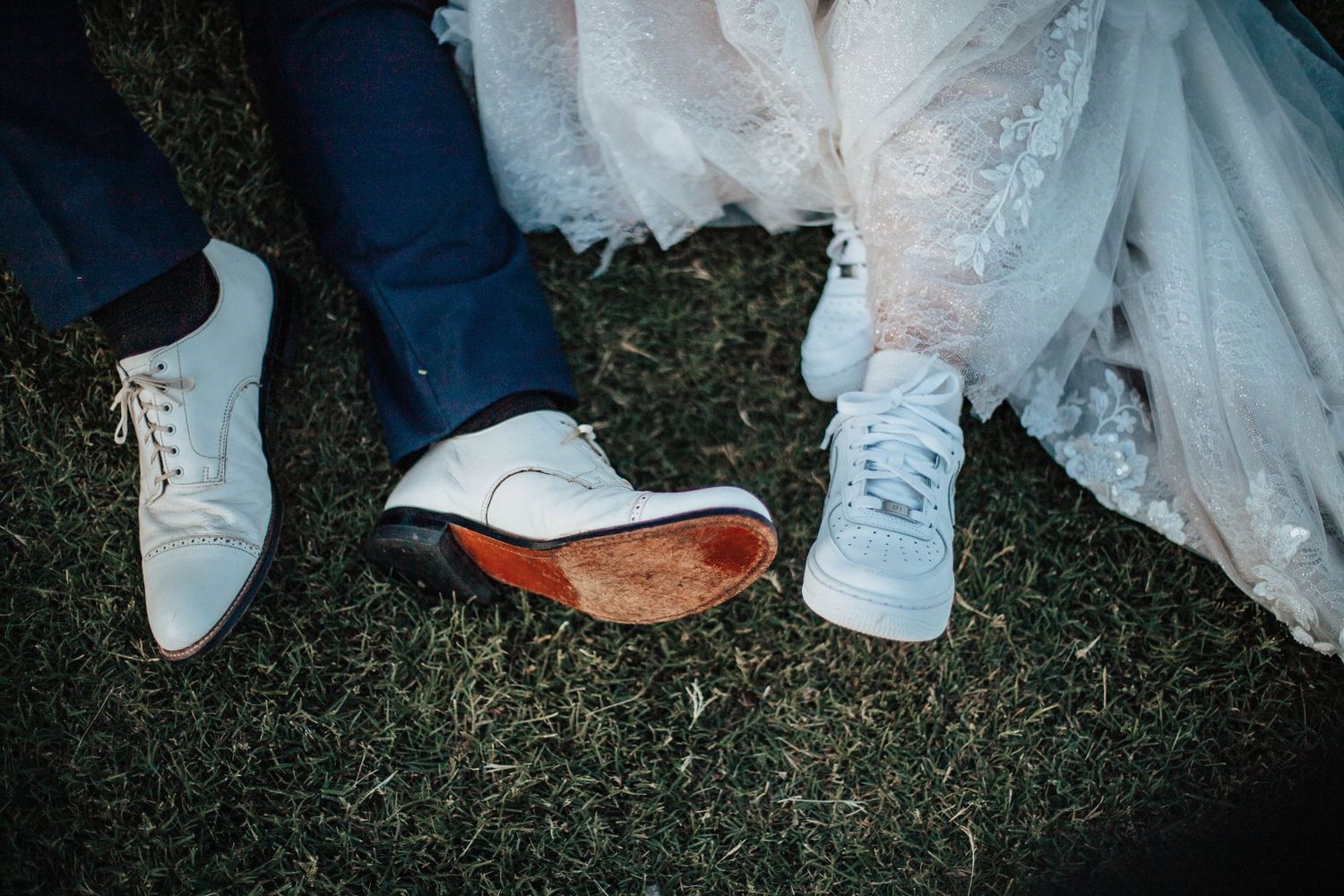 Close-up of wedding couple's shoes on grass, groom in white dress shoes and bride in white sneakers.