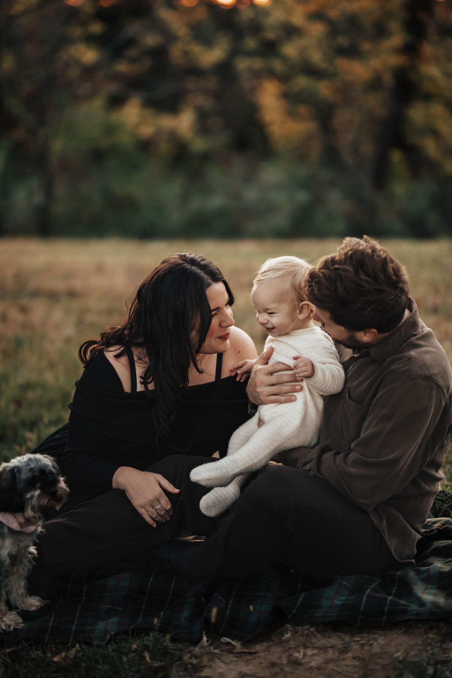 A family sits on a blanket in a park, smiling at their baby in autumn.