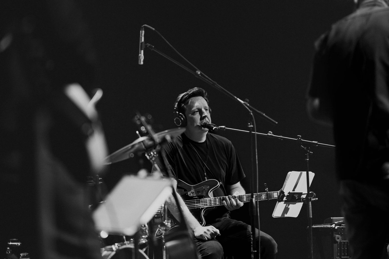 Black and white photo of a guitarist singing into a microphone on stage during a live music performance.