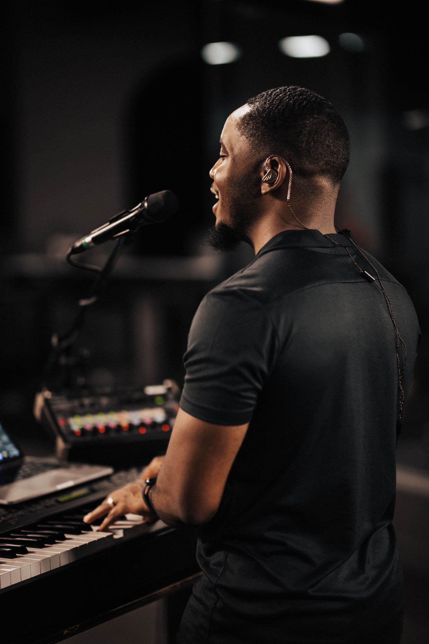 Black man singing into a microphone while playing keyboard in a dark recording studio.