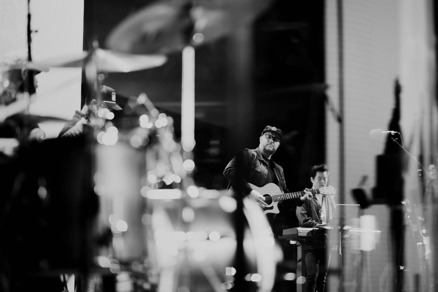 Black and white photo of a guitarist performing on stage with bokeh lights and drum kit visible in background.