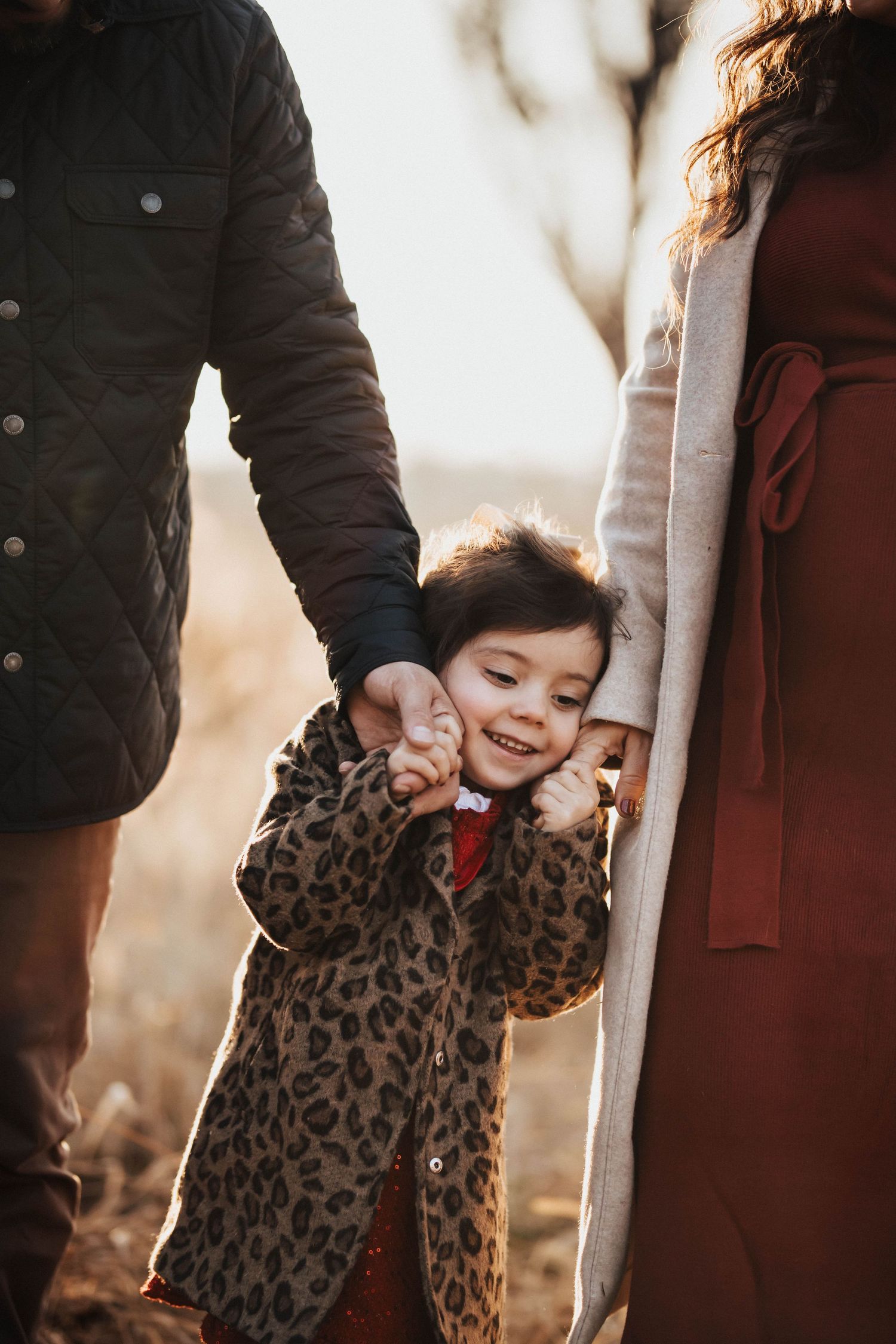 Smiling young girl in leopard coat holds parents' hands outdoors at golden hour.