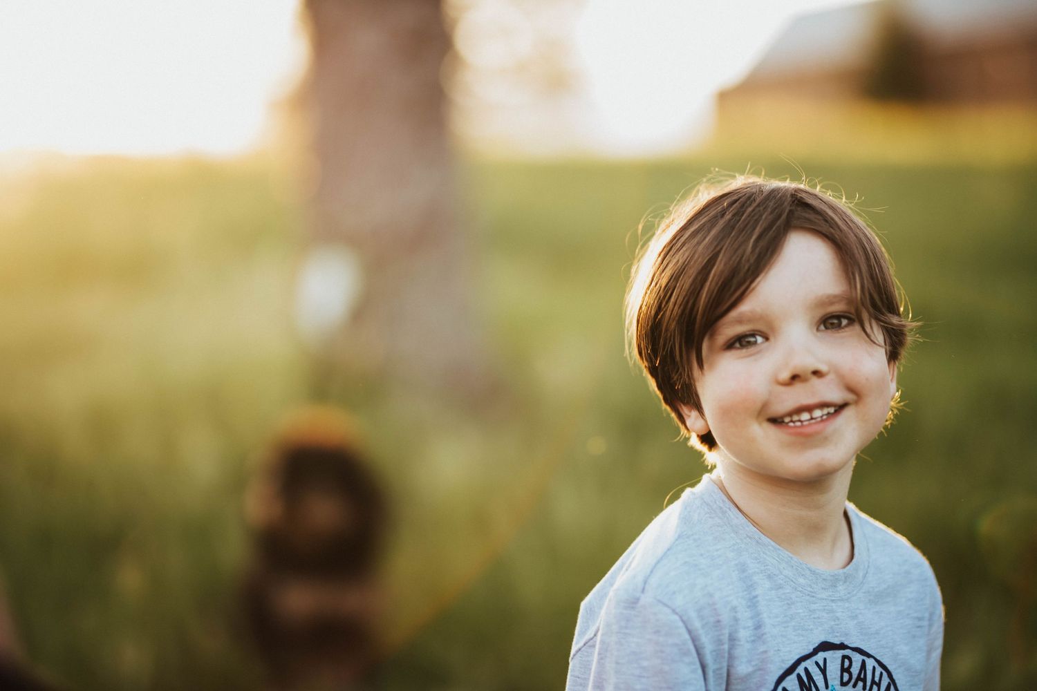 Smiling young boy with brown hair in a gray t-shirt outdoors in a sunny green field at golden hour.