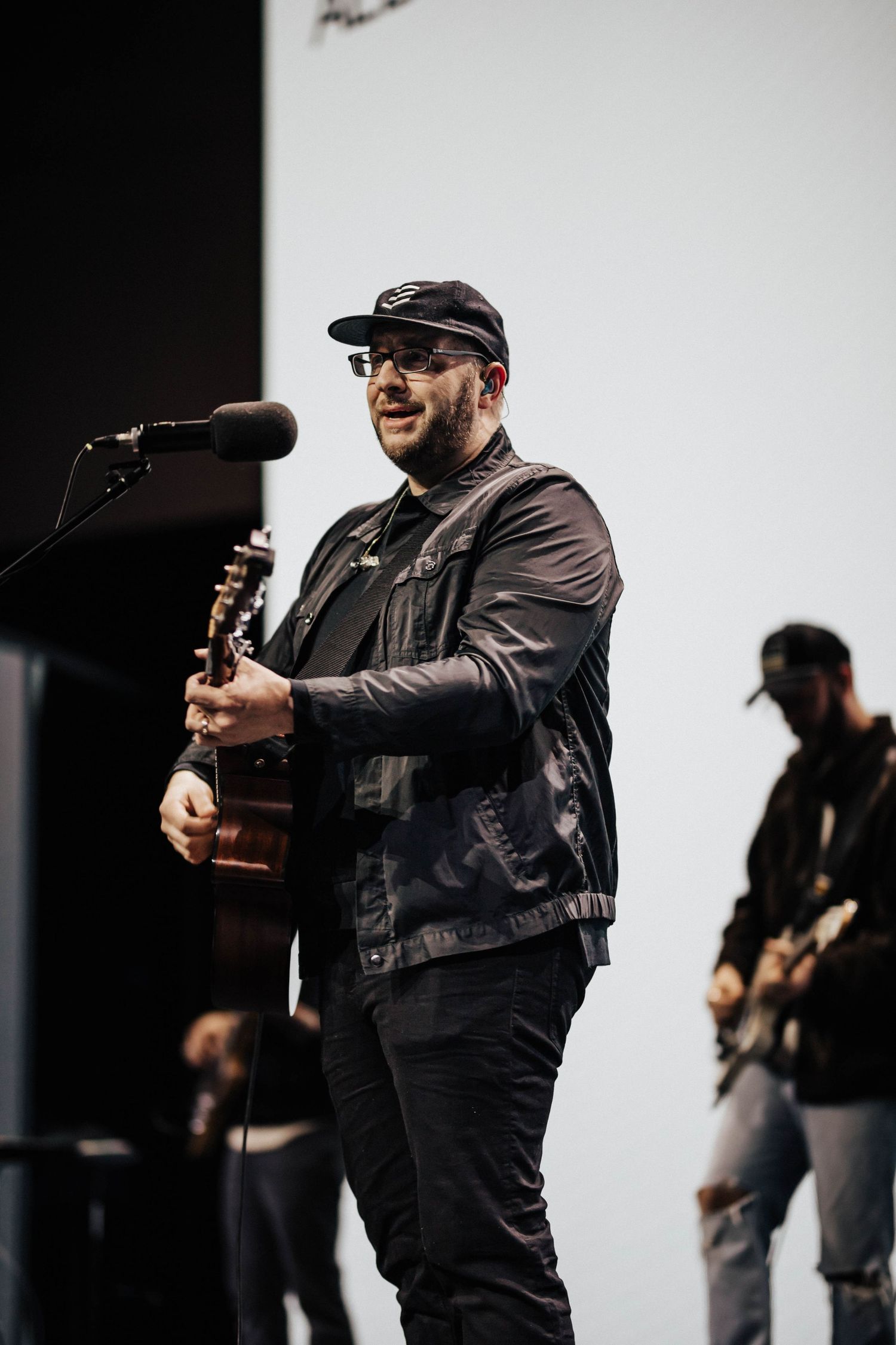 Bearded guitarist in black jacket and cap performs on stage with band members in background.