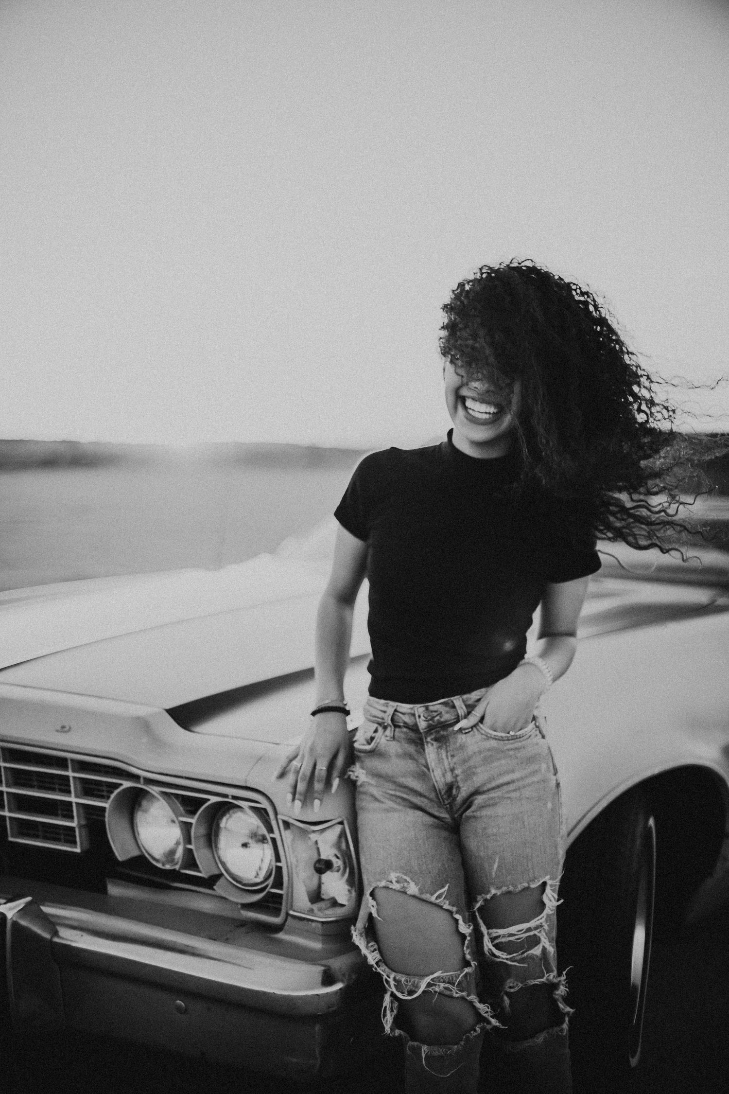 Smiling woman with curly hair leans against vintage car in black and white photo.