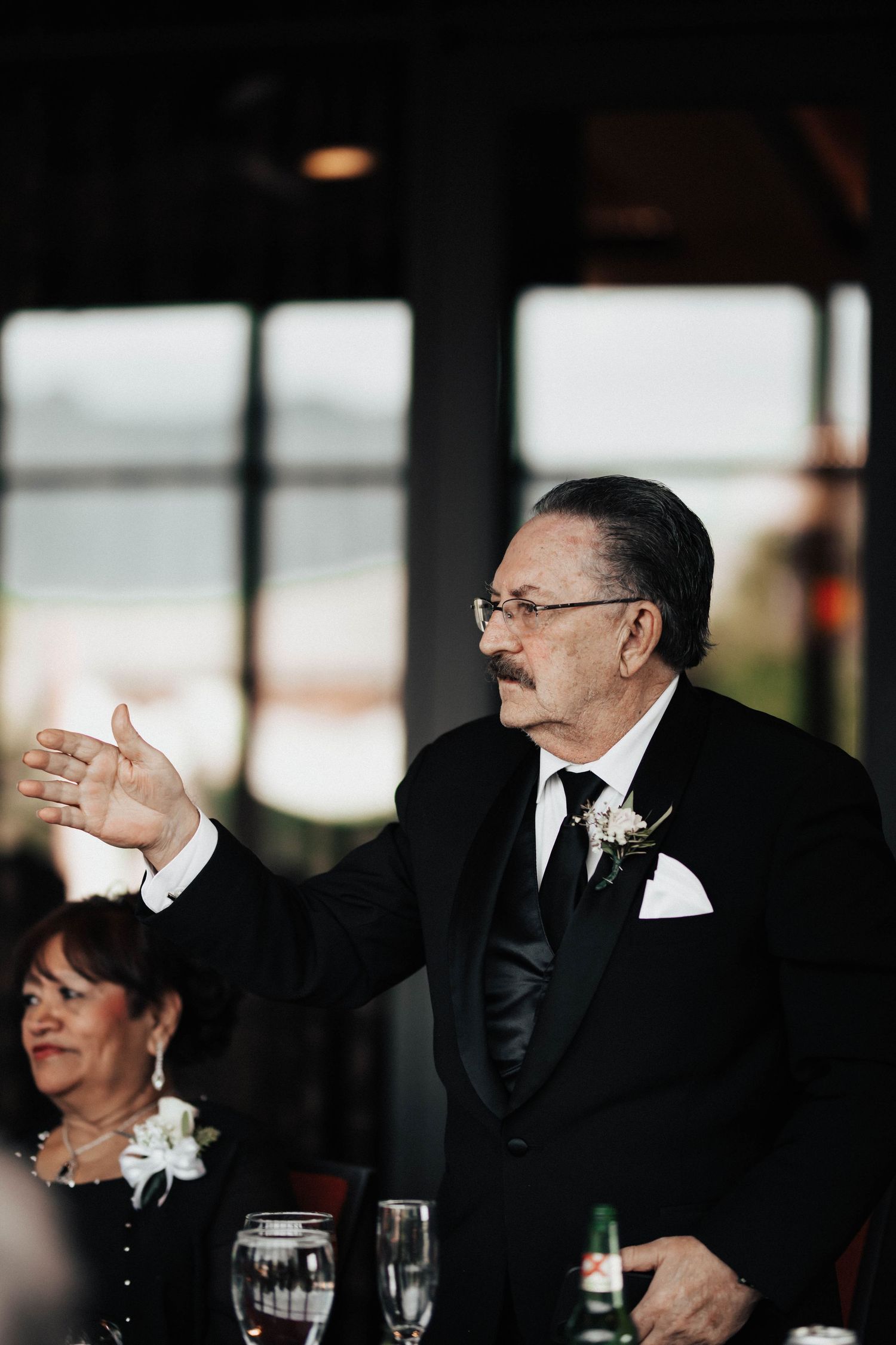 A man in a black tuxedo with a boutonniere gestures while giving a speech at a formal wedding reception.