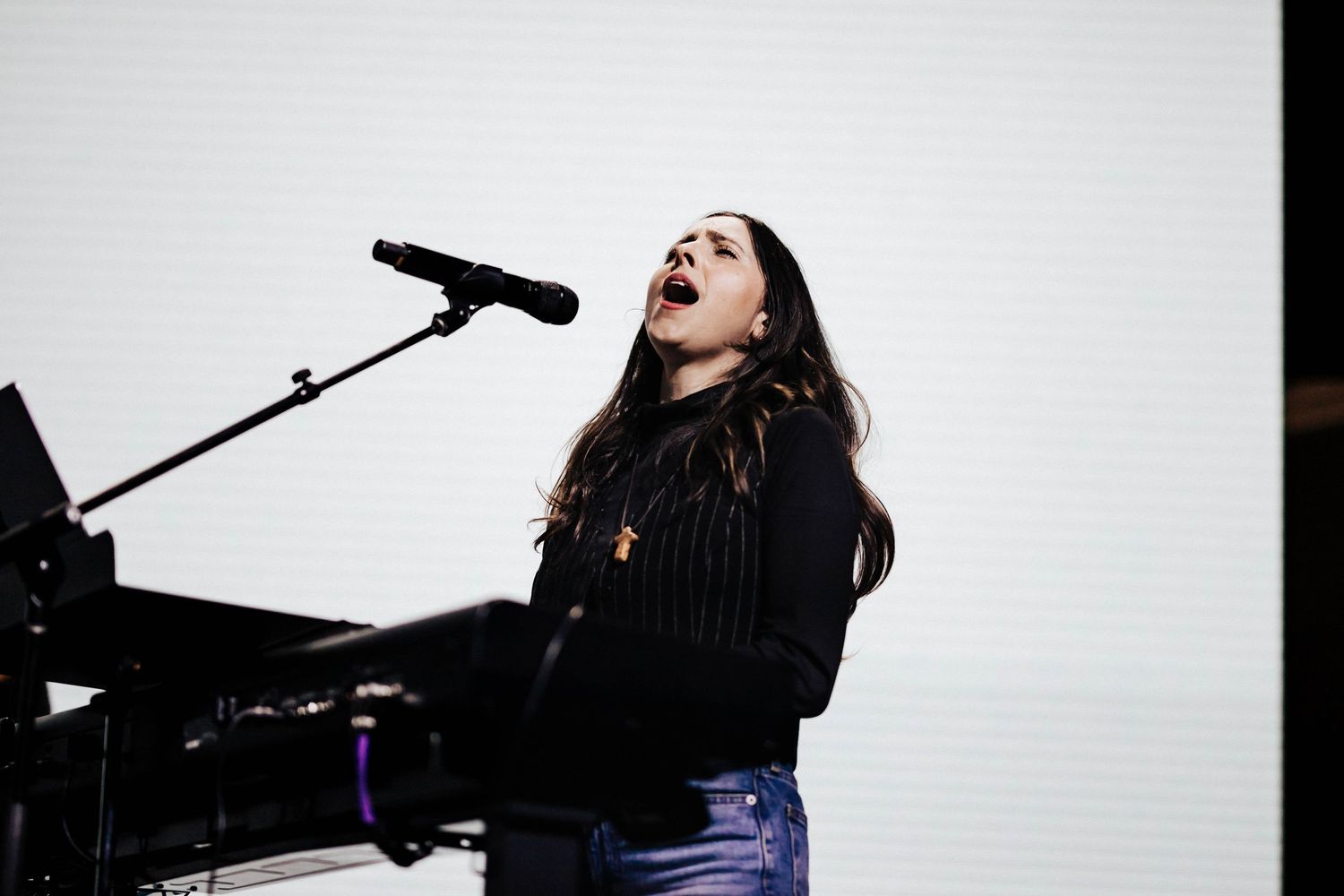 Young female vocalist singing passionately at a piano and microphone on a bright lit stage.