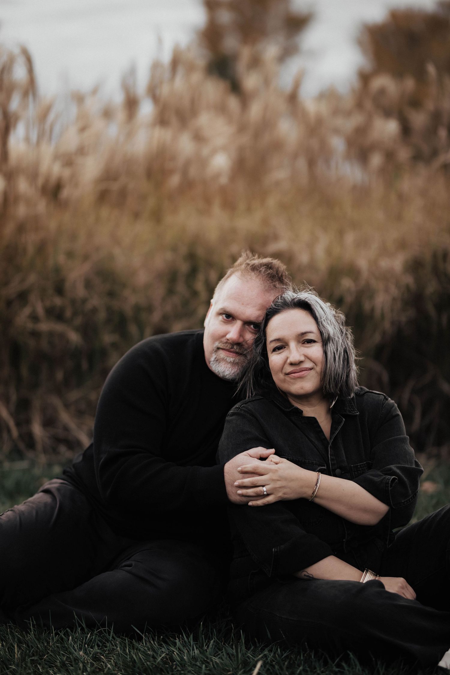Couple sitting together on grass in front of tall autumn reeds, smiling warmly at camera.