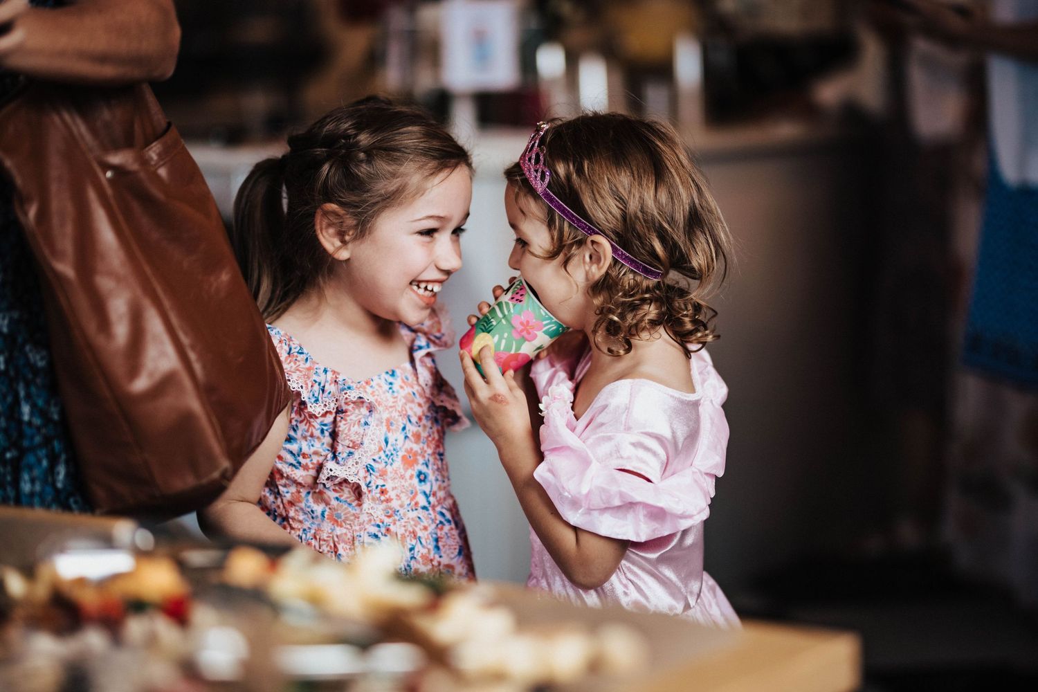 Two young girls in floral dresses share a playful moment eating treats at a party.