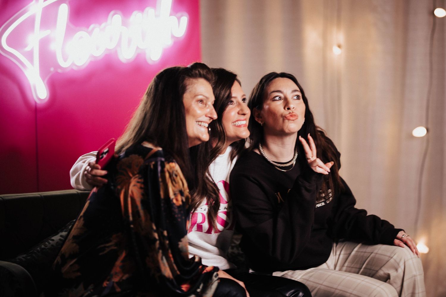 Three women pose together smiling in front of a pink Flourish neon sign.