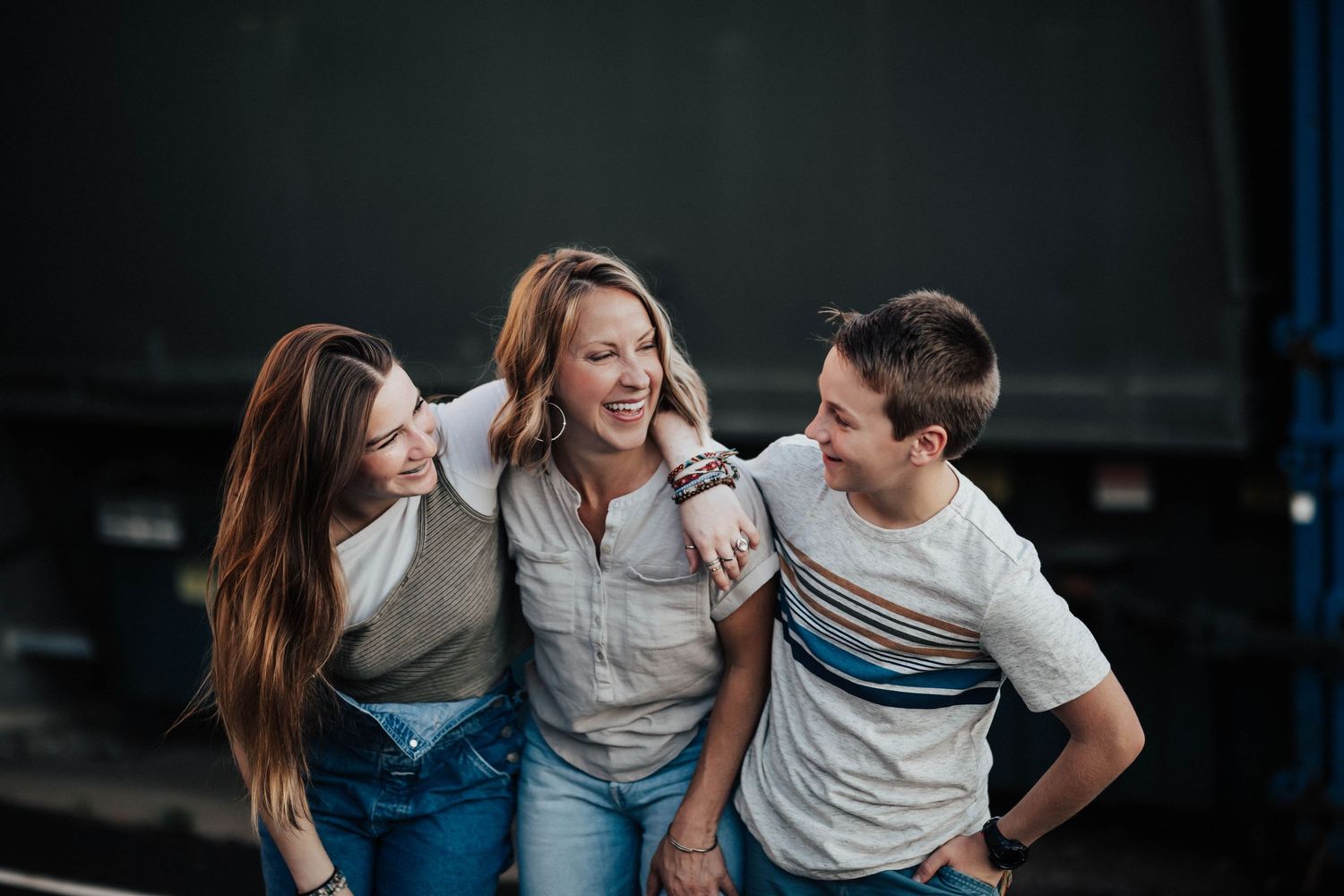 A woman laughs with her teenage daughter and son in a casual outdoor portrait.
