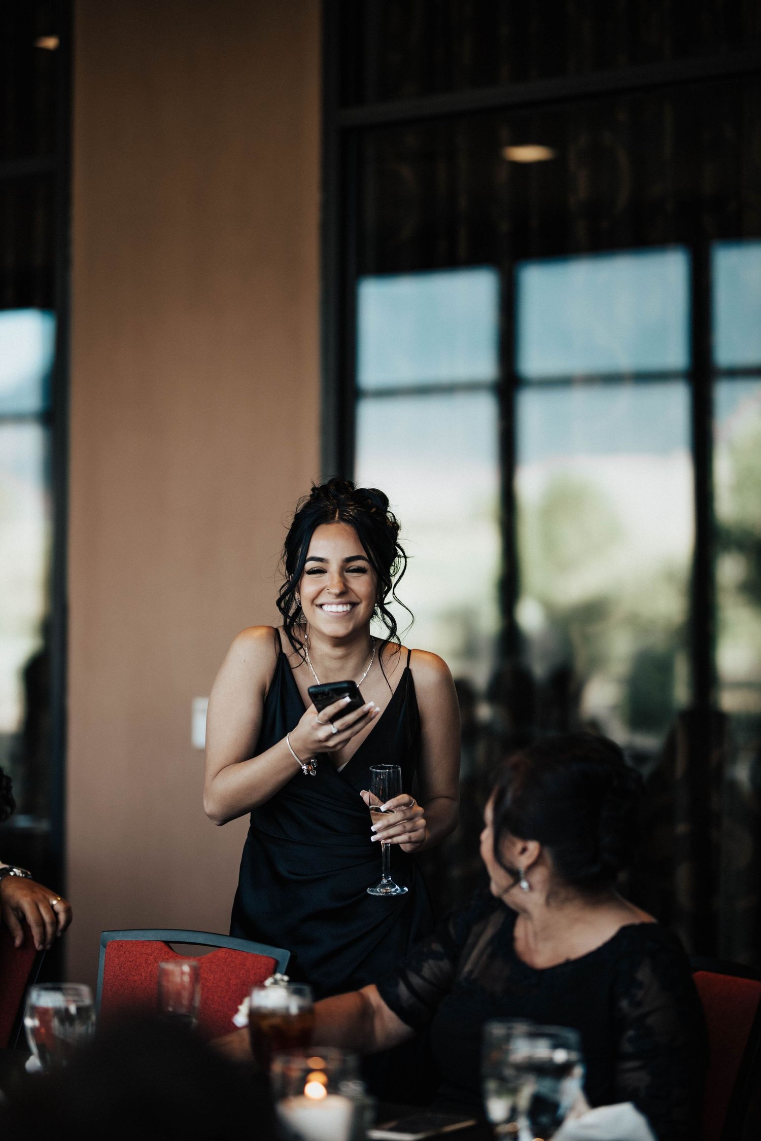 Smiling woman in black dress holding phone and wine glass at elegant indoor event with candles.