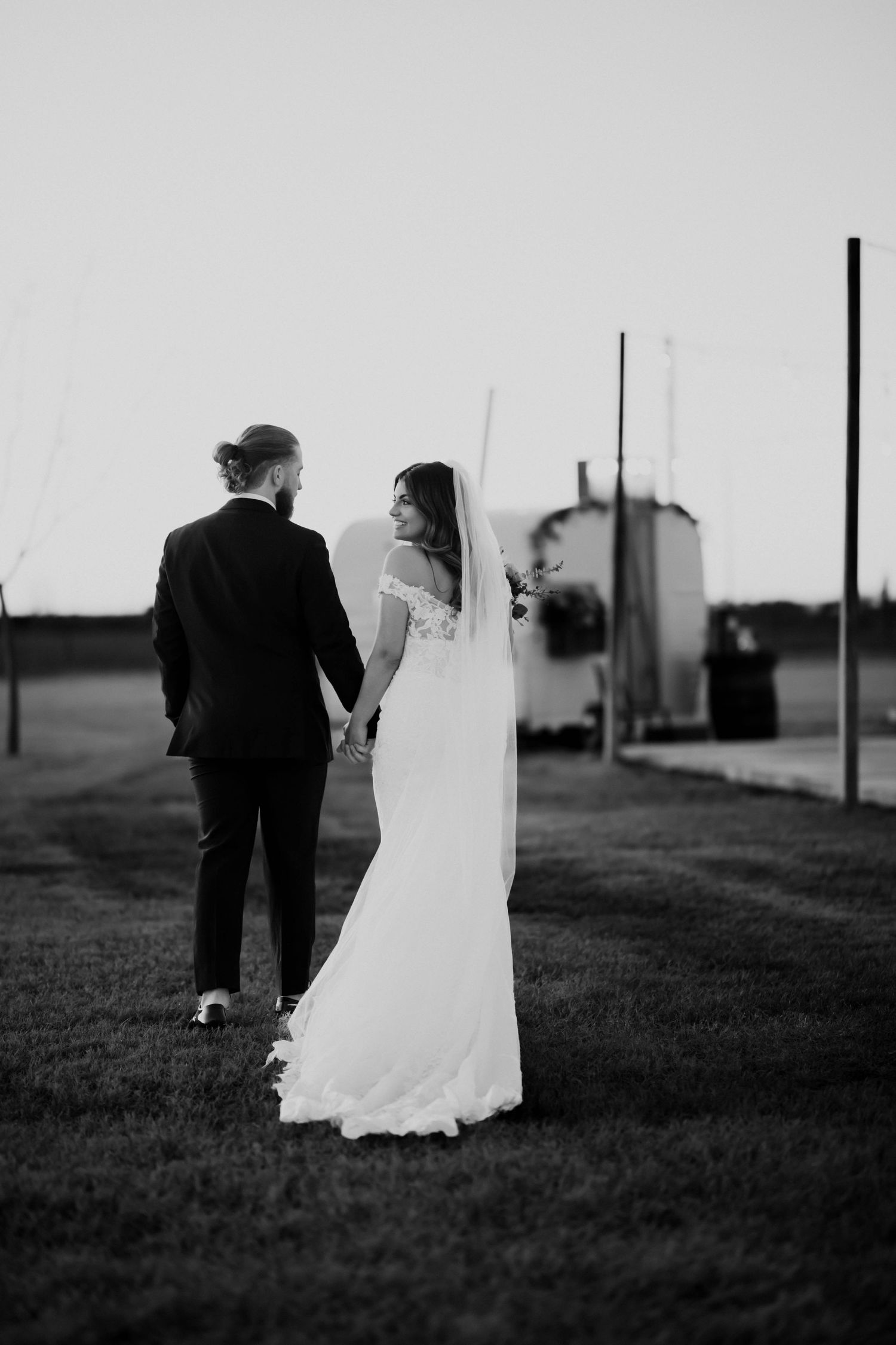 Black and white photo of a bride and groom holding hands walking across a grass field at dusk.