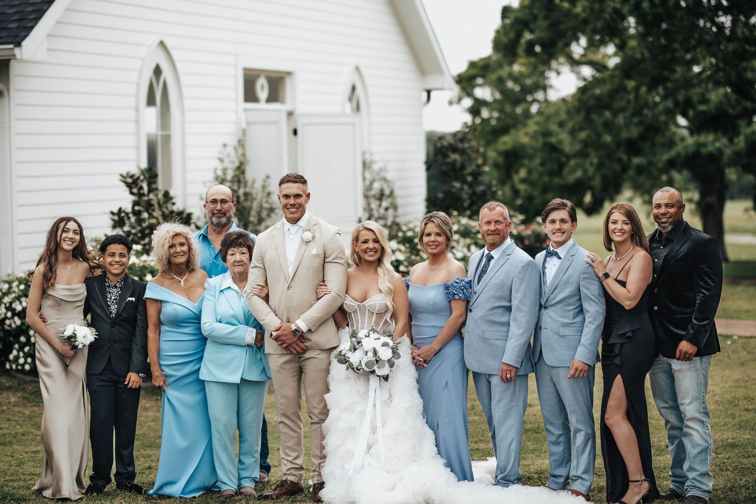 Wedding party posing in blue and beige attire outside a white chapel on a sunny day.
