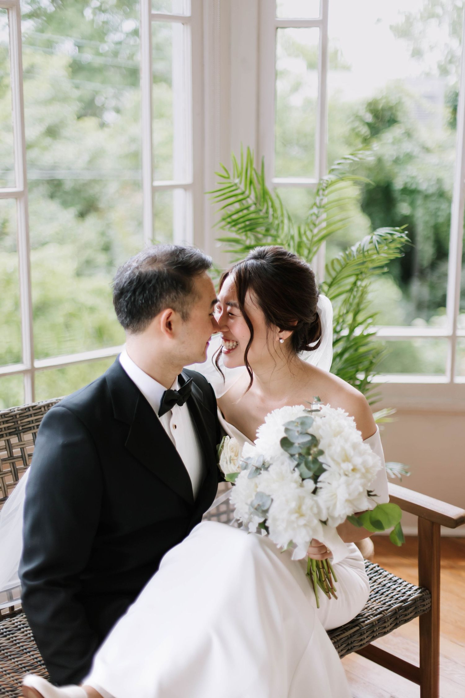 A bride and groom share a romantic moment seated near large windows, bride holding white hydrangea bouquet.