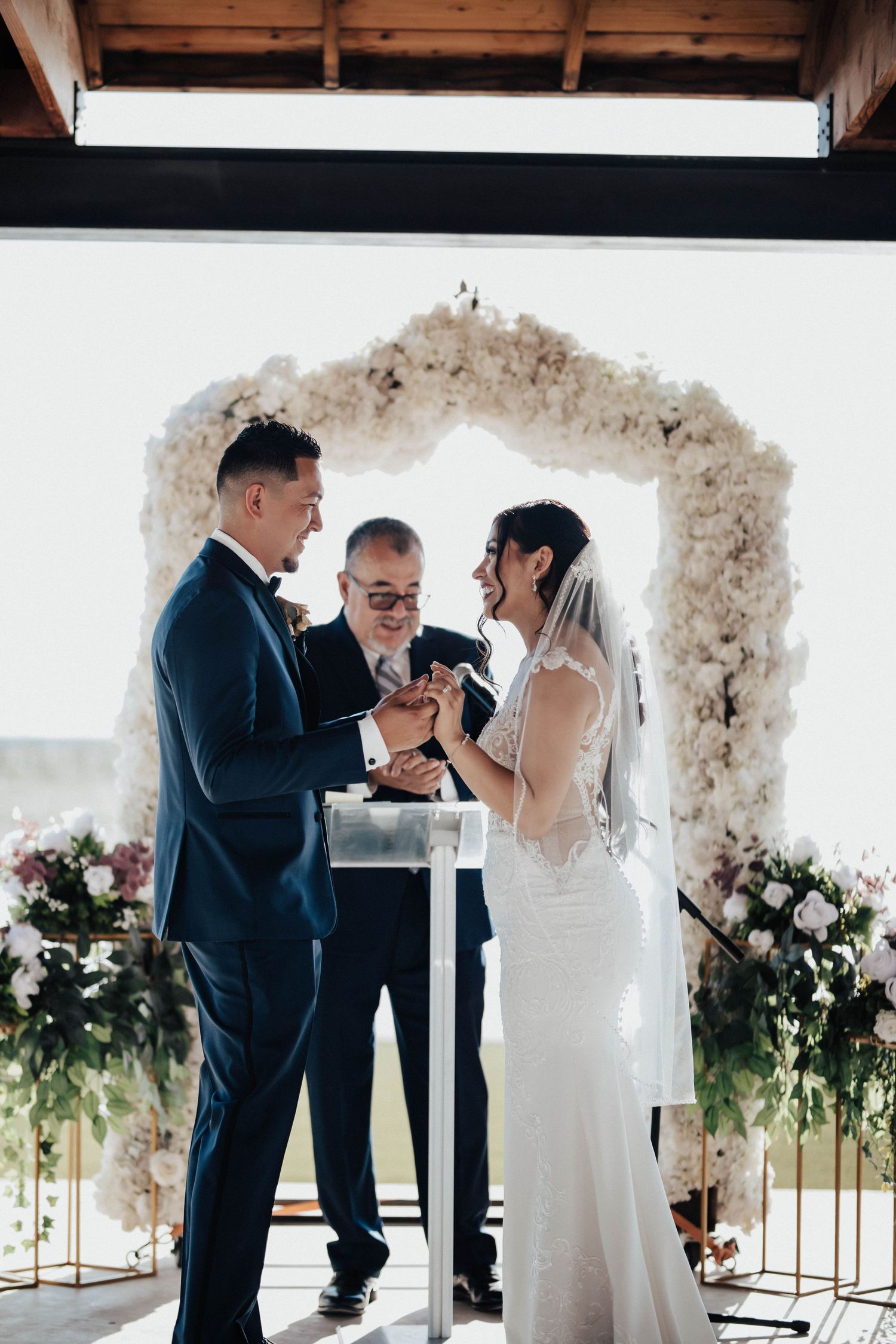 Bride and groom exchange rings at wedding altar adorned with white floral arch, officiant smiling behind them.