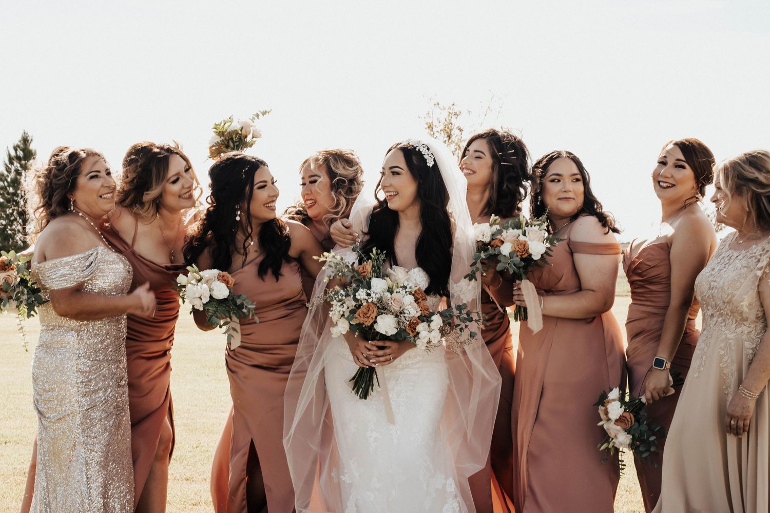 Bride in white gown with bridesmaids in dusty rose dresses holding bouquets, laughing outdoors at wedding.