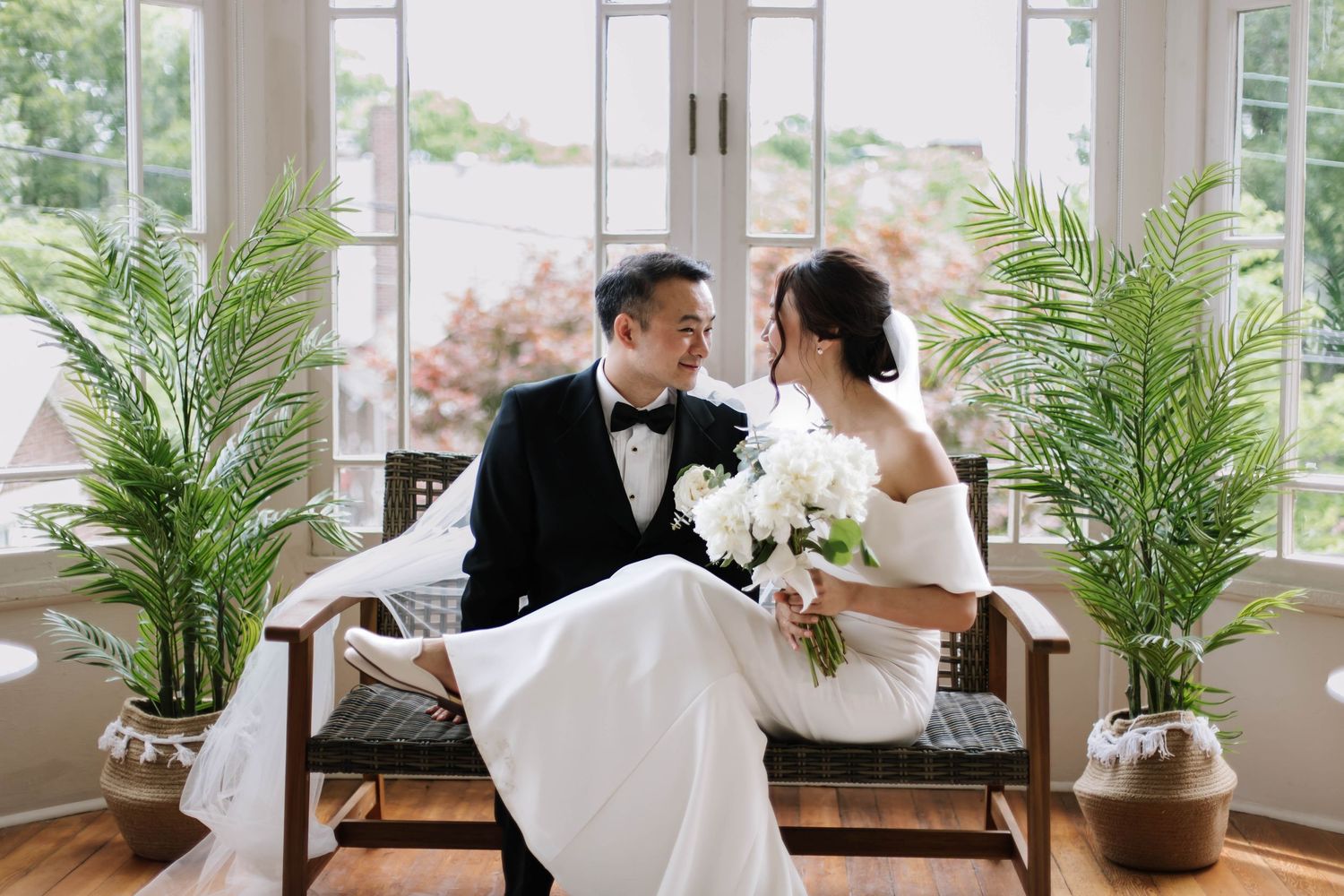 Bride and groom in wedding attire sit on a bench surrounded by palm plants near bright windows.
