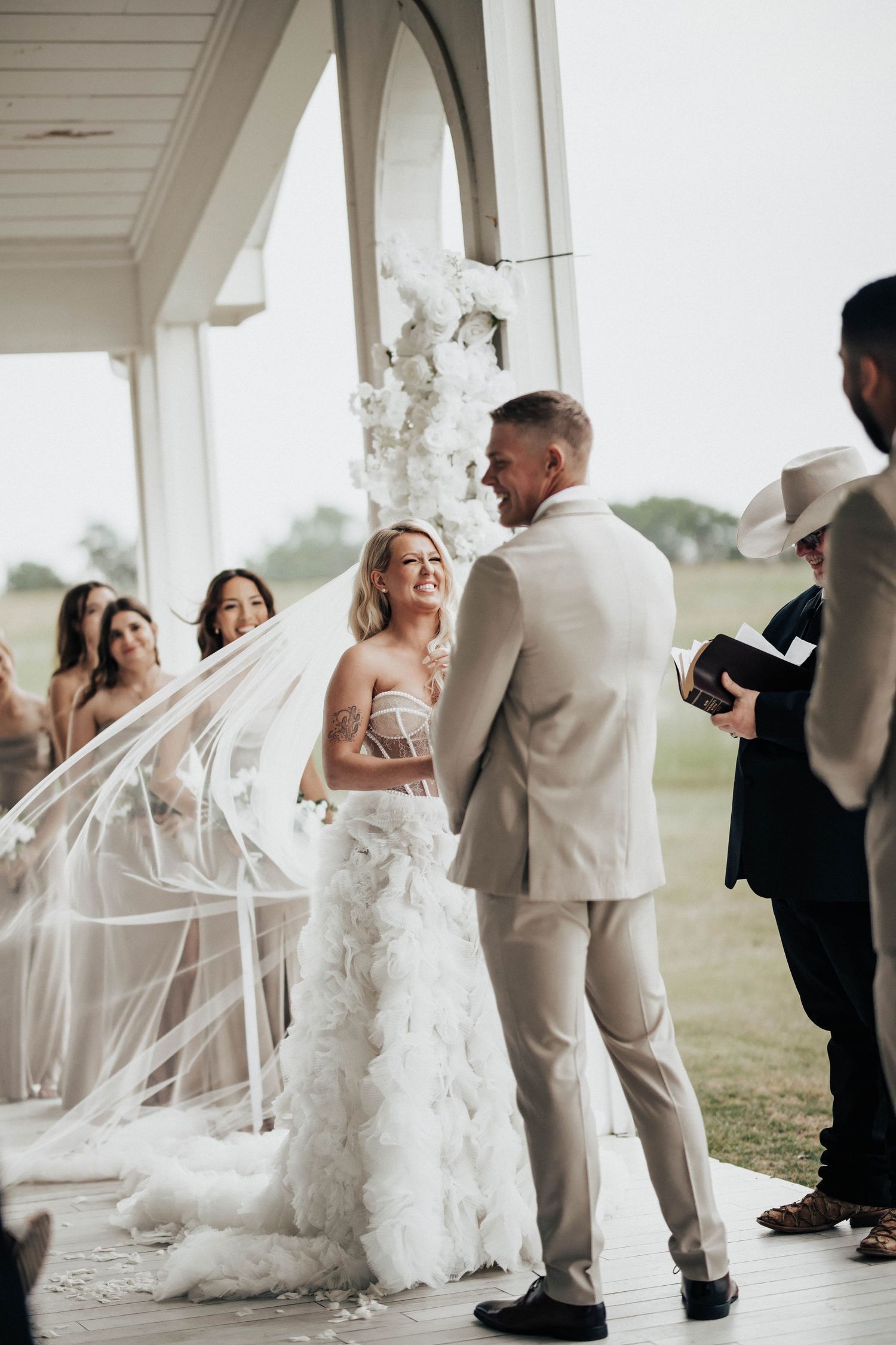 Bride and groom exchange vows at outdoor wedding ceremony, bride wearing ruffled gown with flowing veil.