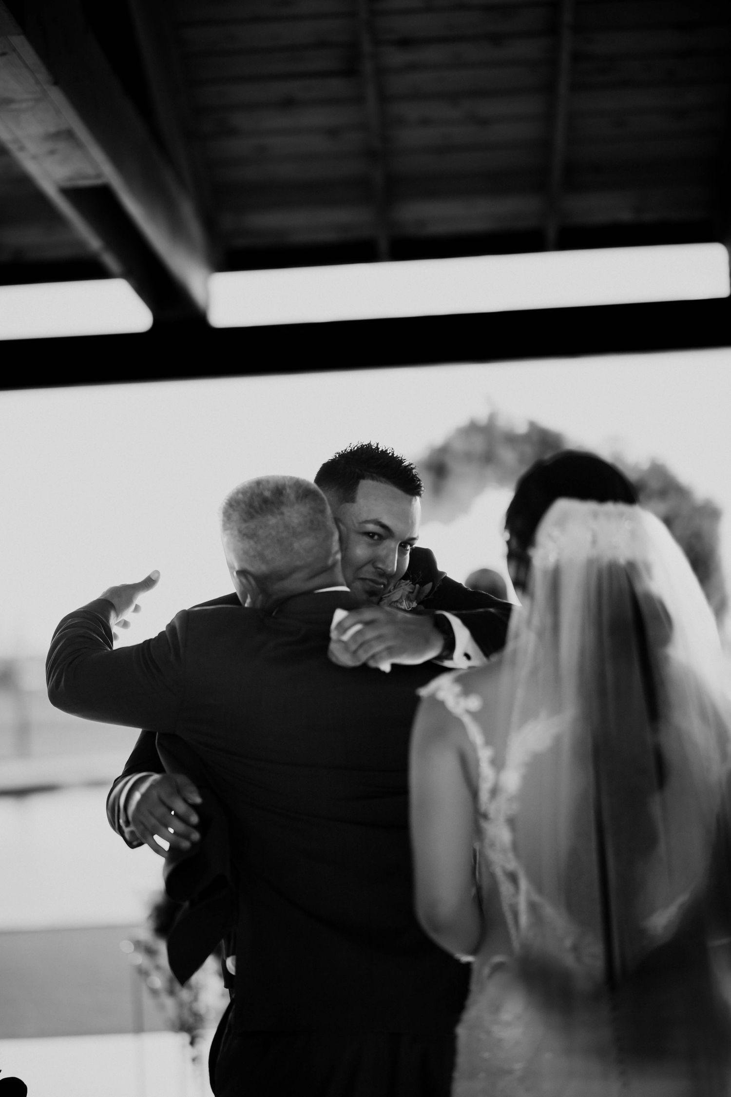 Black and white photo of groom embracing older man at wedding ceremony, bride in veil visible in foreground.