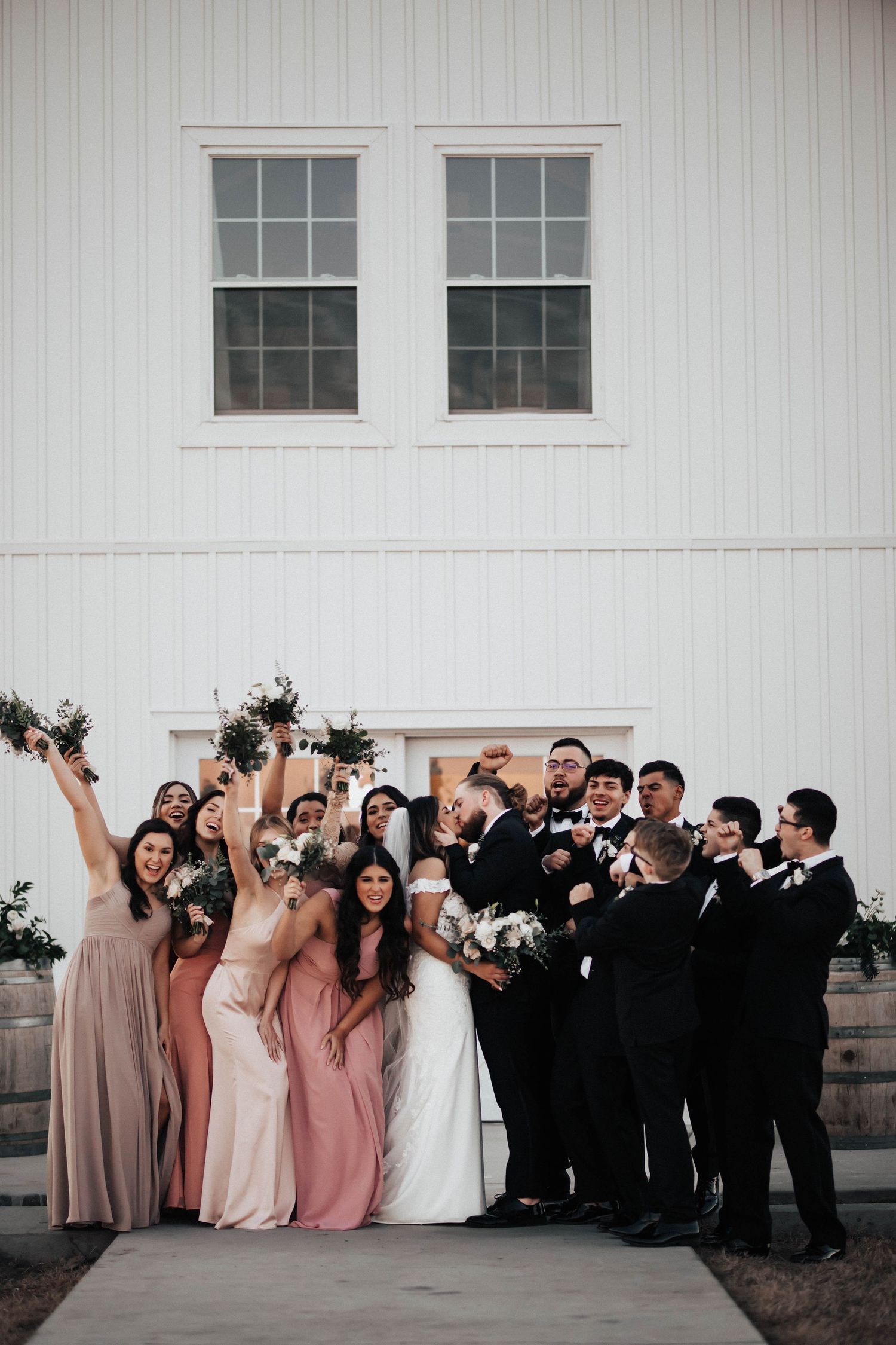 Wedding party celebrating in front of white barn, bridesmaids in blush gowns and groomsmen in black tuxedos cheering.