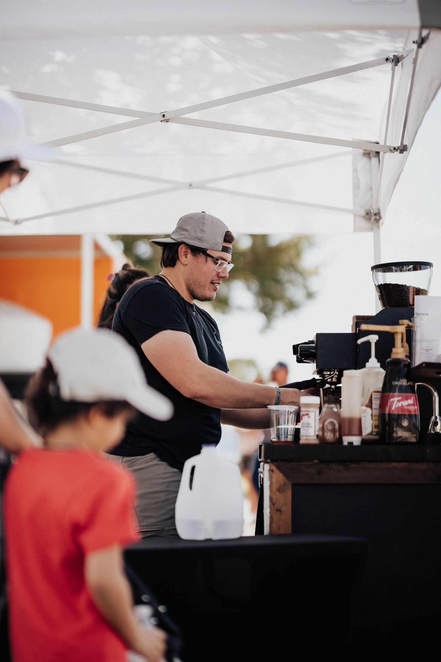 Barista in cap prepares coffee drinks at outdoor espresso stand under white canopy.