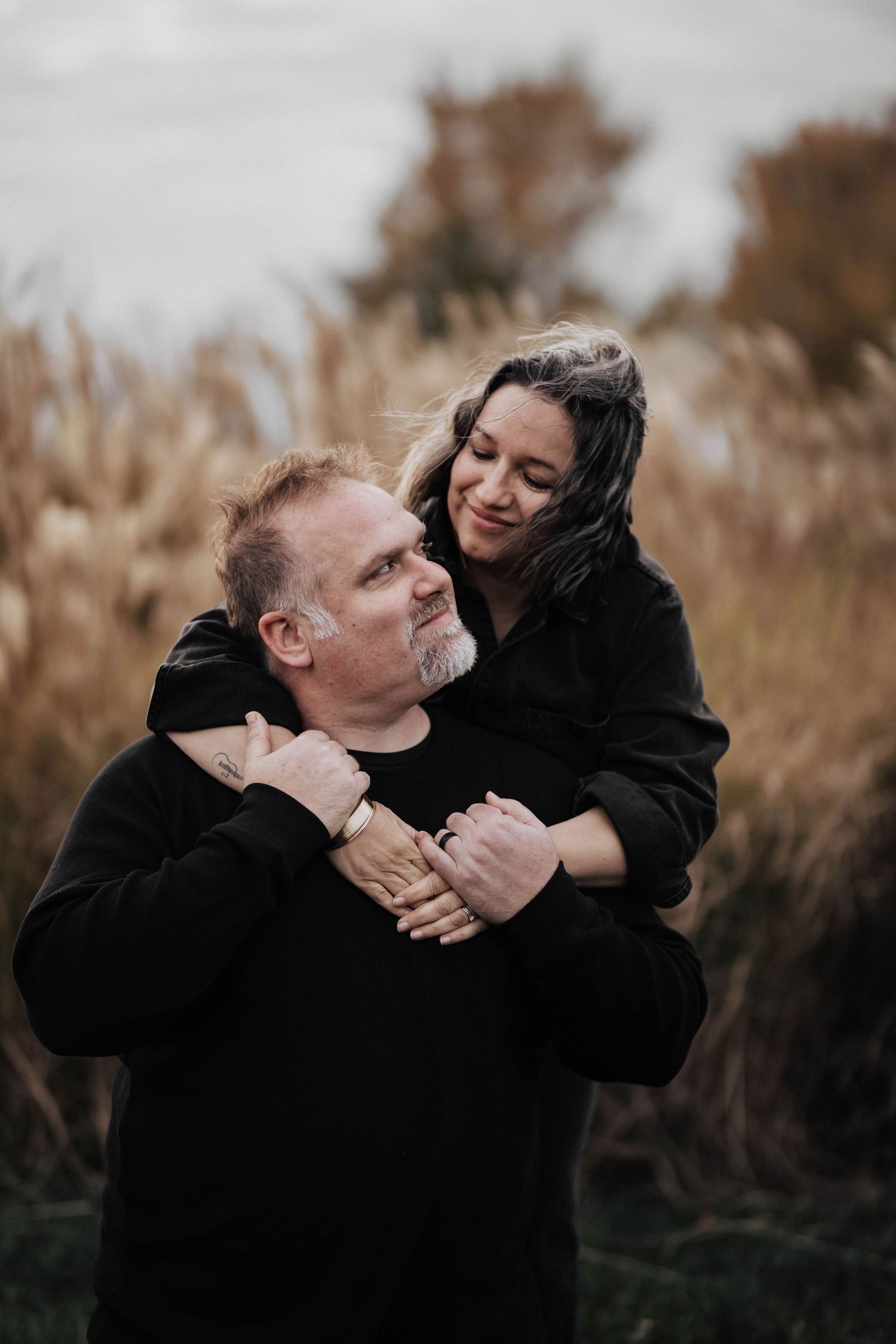 Couple embracing outdoors in autumn field, woman hugging man from behind among tall golden grasses.