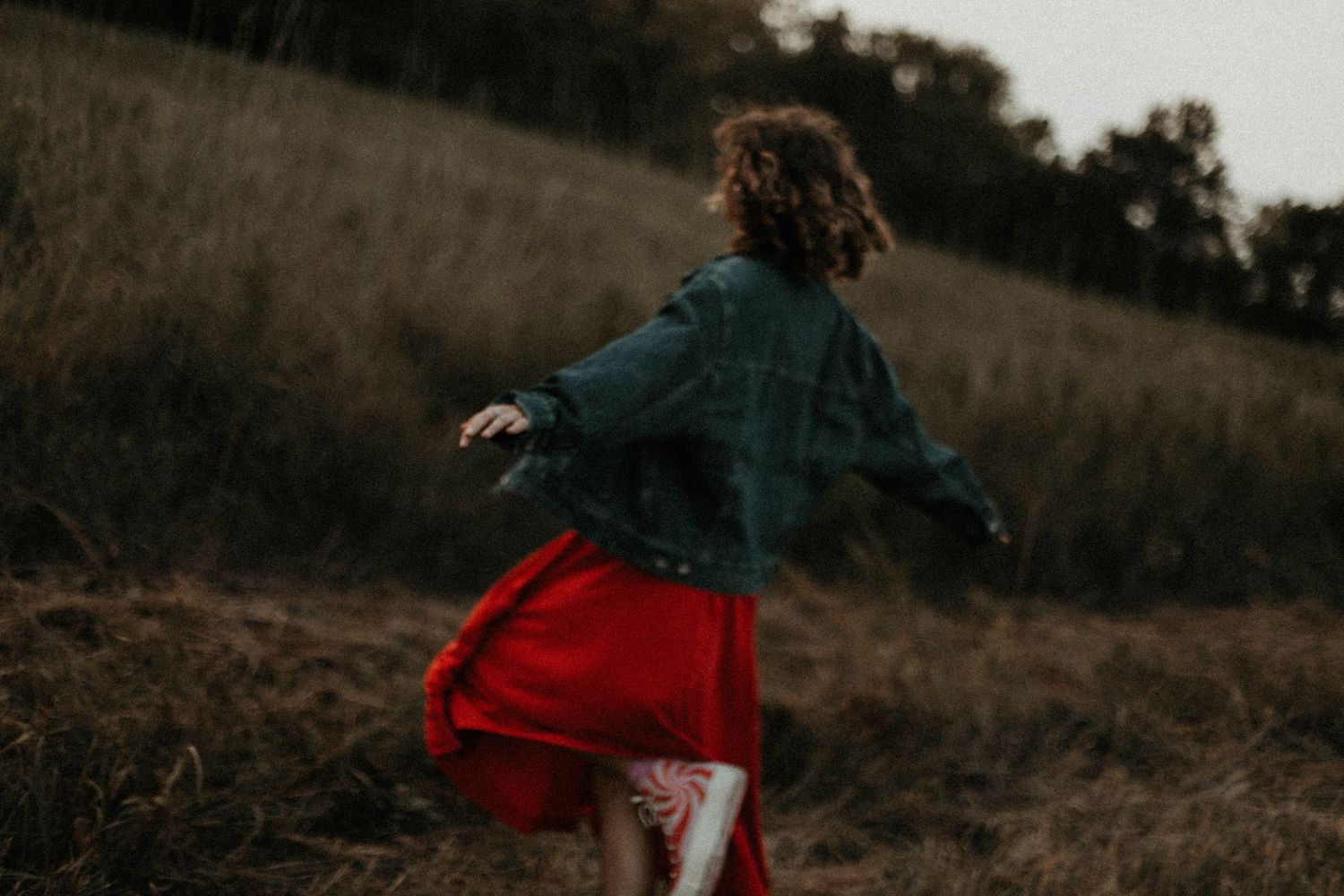 A young girl in a red skirt and denim jacket twirls joyfully in a golden field at dusk.