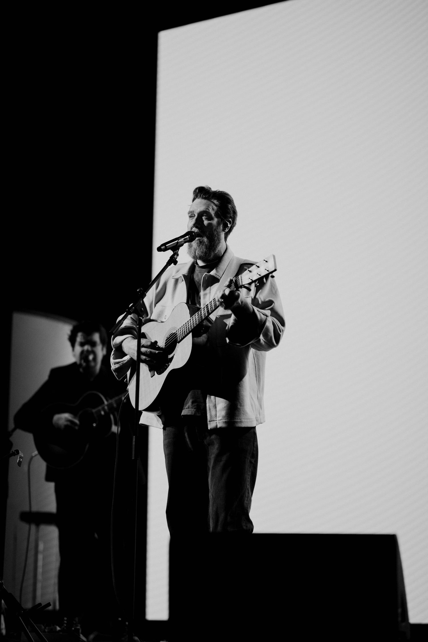 Black and white photo of bearded guitarist singing at microphone on stage with large white screen behind.