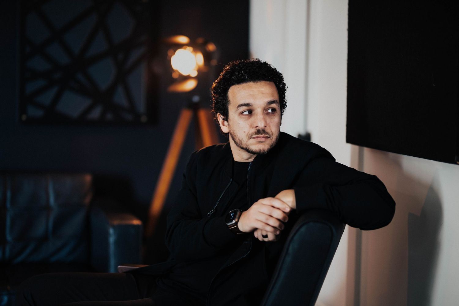 Young man in black outfit sitting pensively in a modern dark interior with ambient lighting.