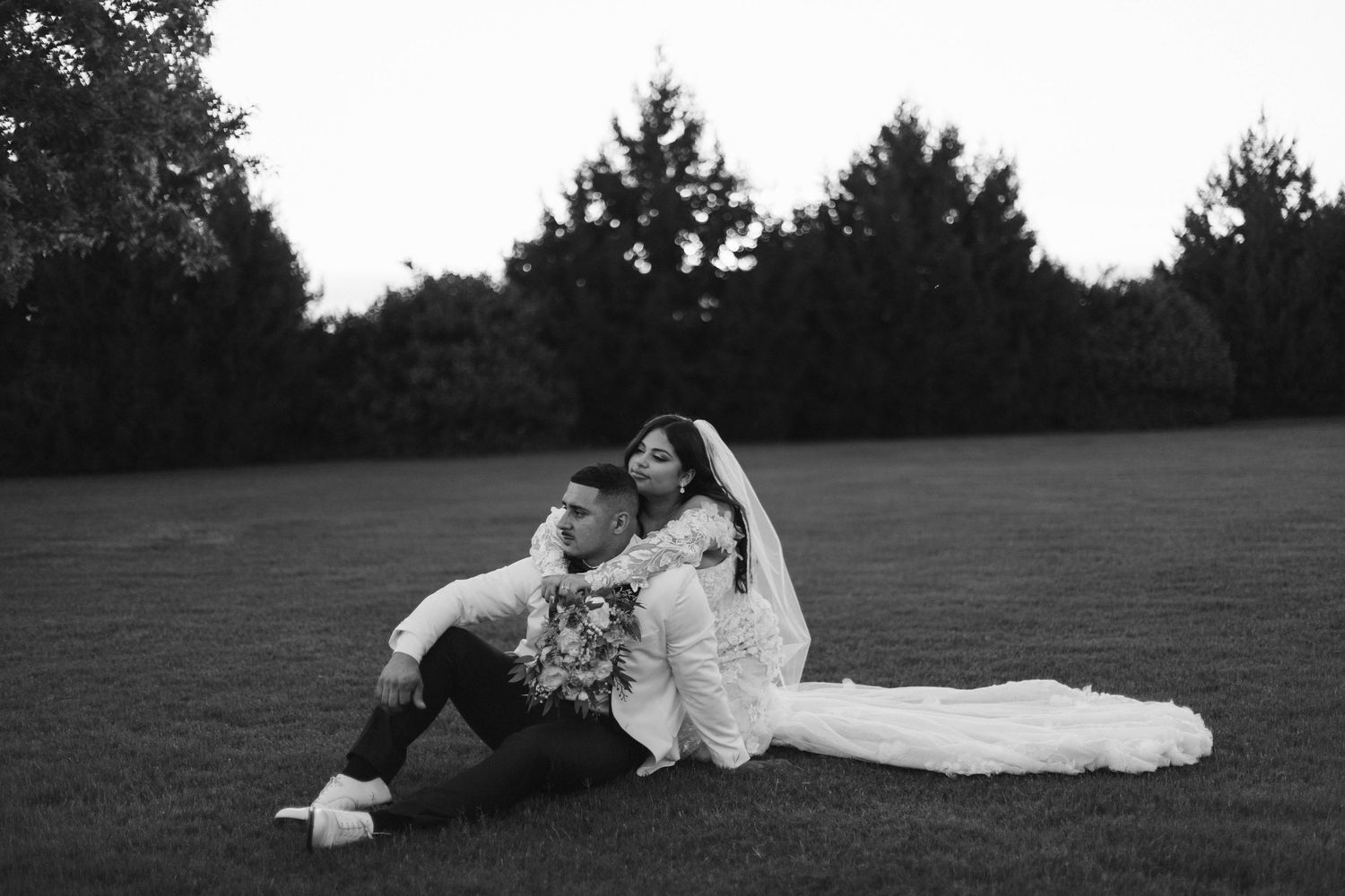 Black and white photo of bride and groom sitting together on grass at dusk in a romantic outdoor wedding portrait.