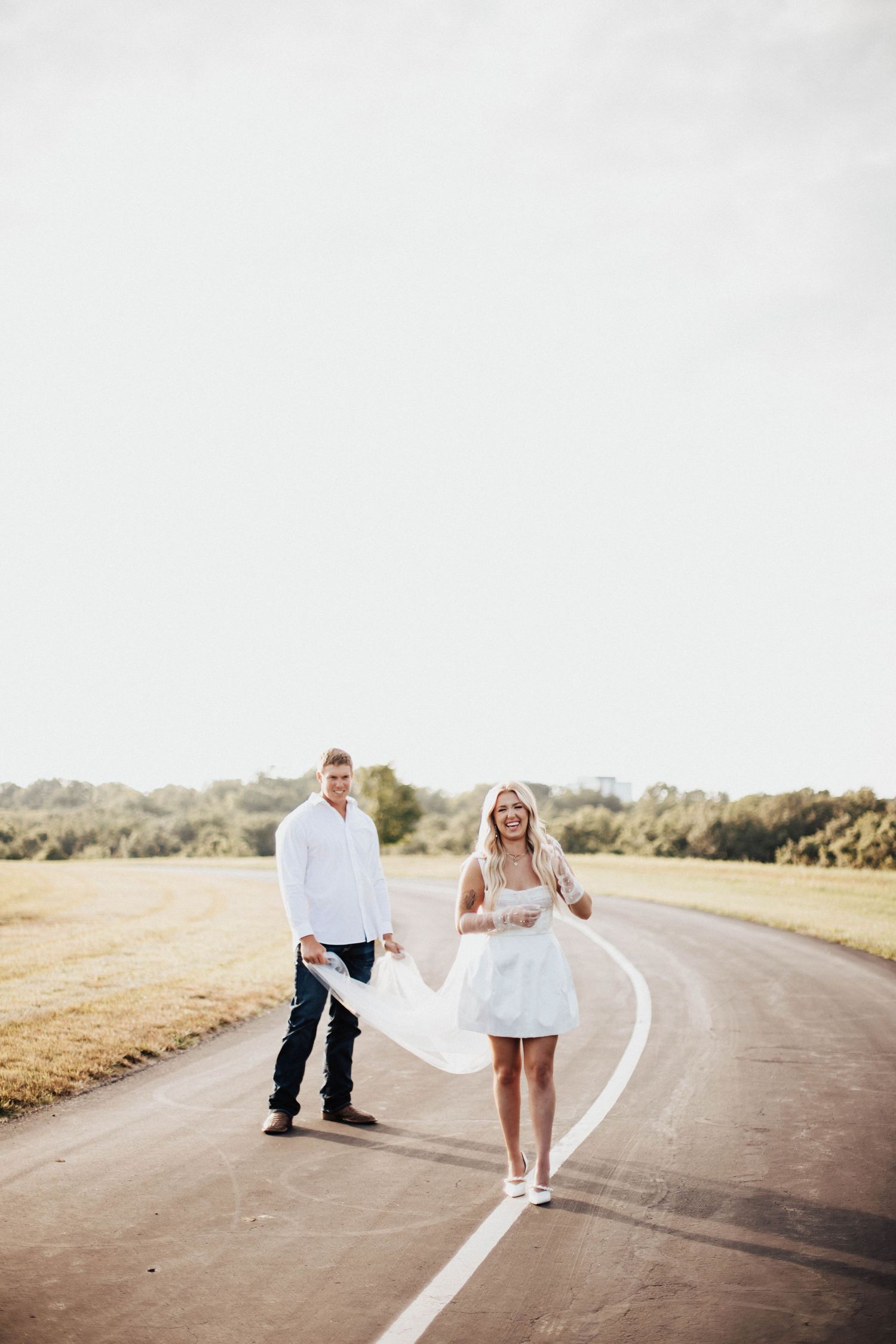 Couple in white outfits walking on a rural road, woman in mini dress holding fabric, man in jeans smiling behind her.