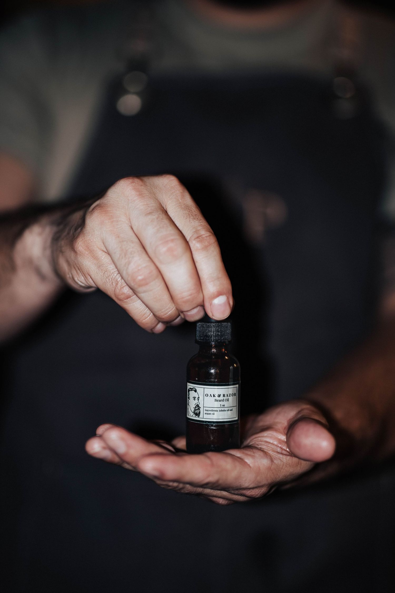 Barber in apron carefully dispensing beard oil from a small dark glass bottle.