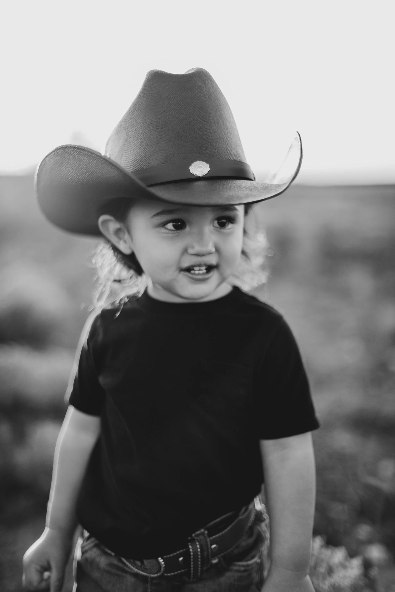 Black and white portrait of toddler wearing oversized cowboy hat and black shirt outdoors.