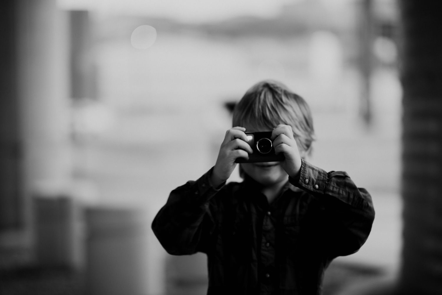 Black and white photo of a child holding a camera up to their face, taking a picture outdoors.