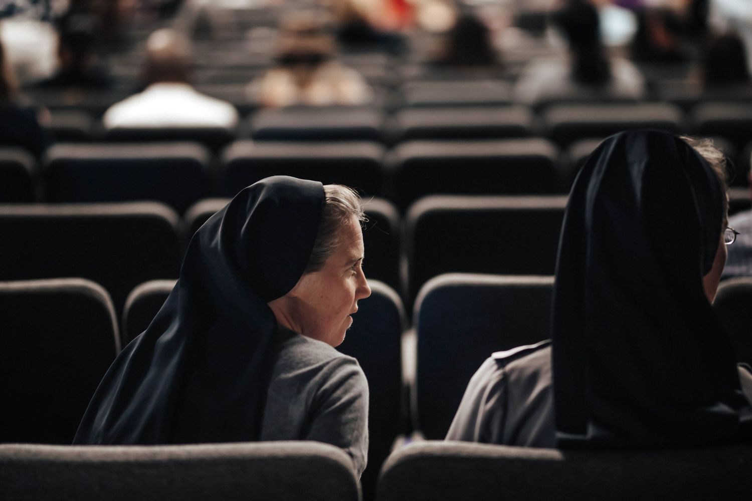Two nuns wearing black habits sit together in auditorium seats during a religious gathering.