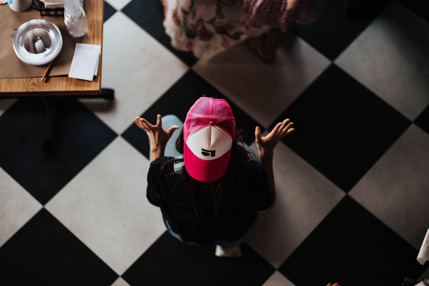 Overhead view of person wearing pink cap sitting at checkered floor table with hands raised.
