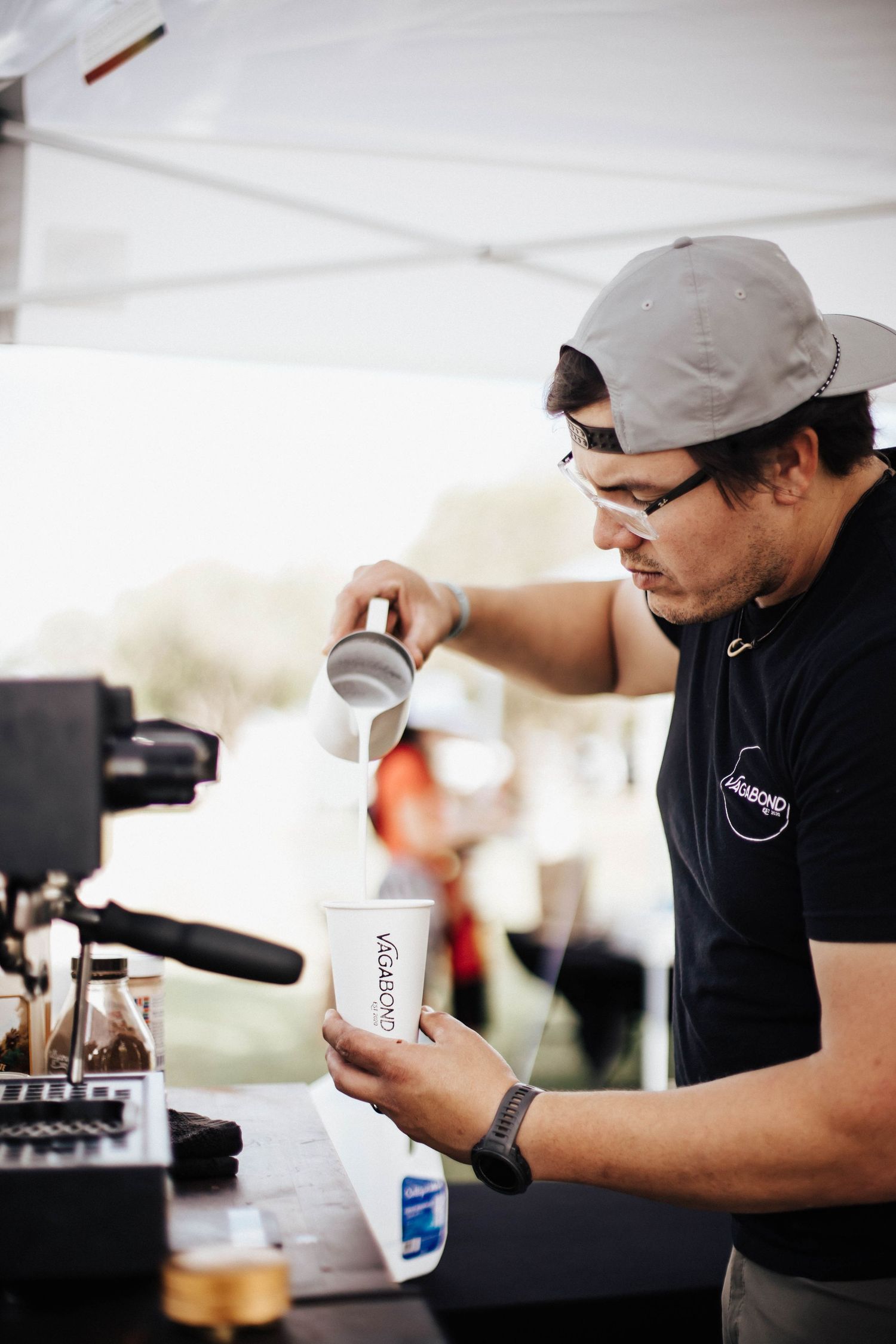 Barista in black cap pours milk into Vagabond Coffee cup at outdoor espresso stand.