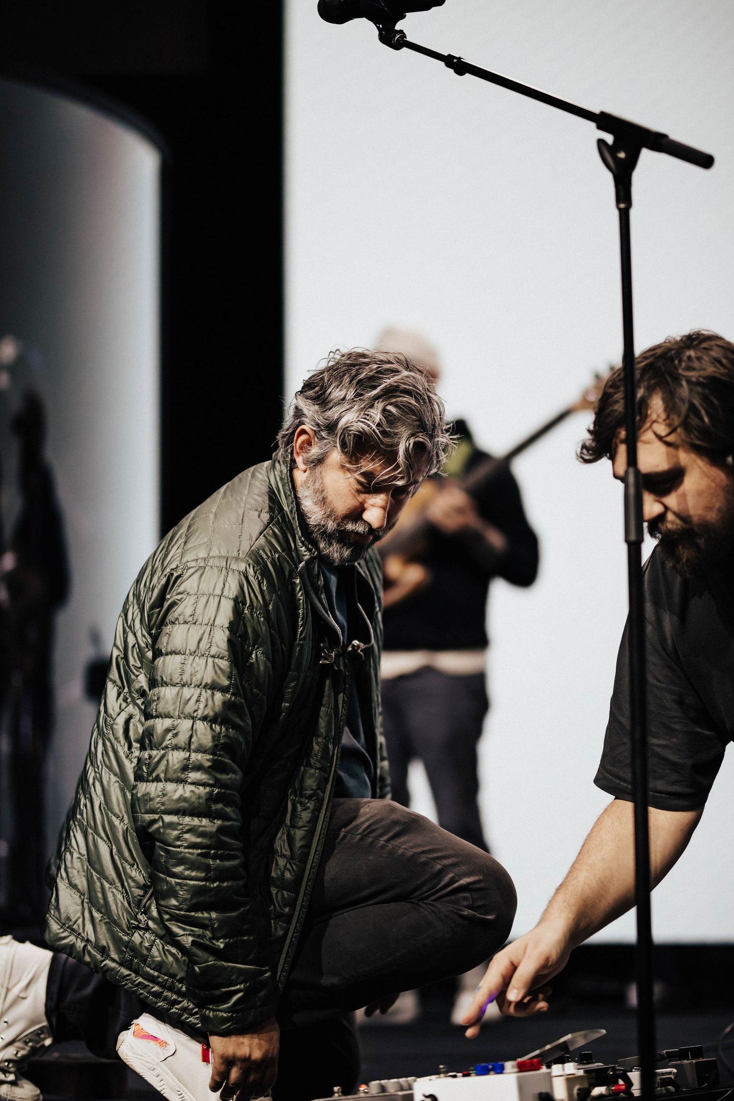 Bearded man in green jacket crouching on stage adjusting audio equipment during a live performance.
