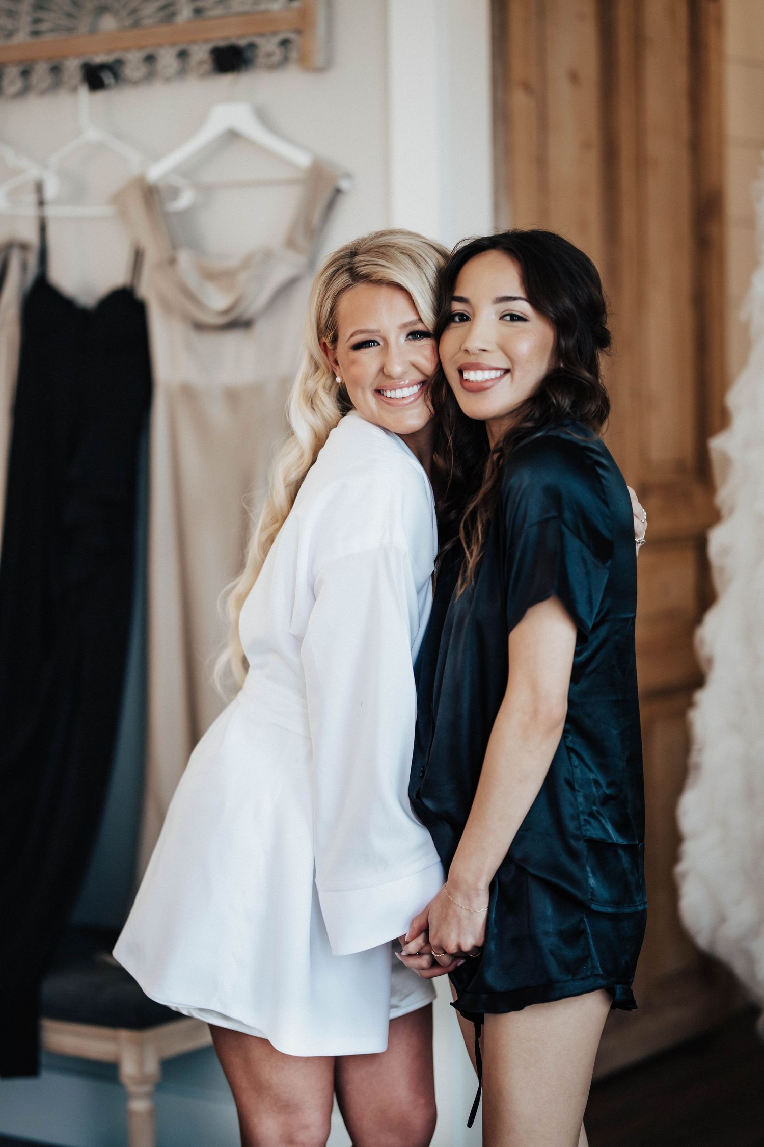 Two smiling women in silk robes pose together in a bridal getting-ready room with dresses hanging in background.