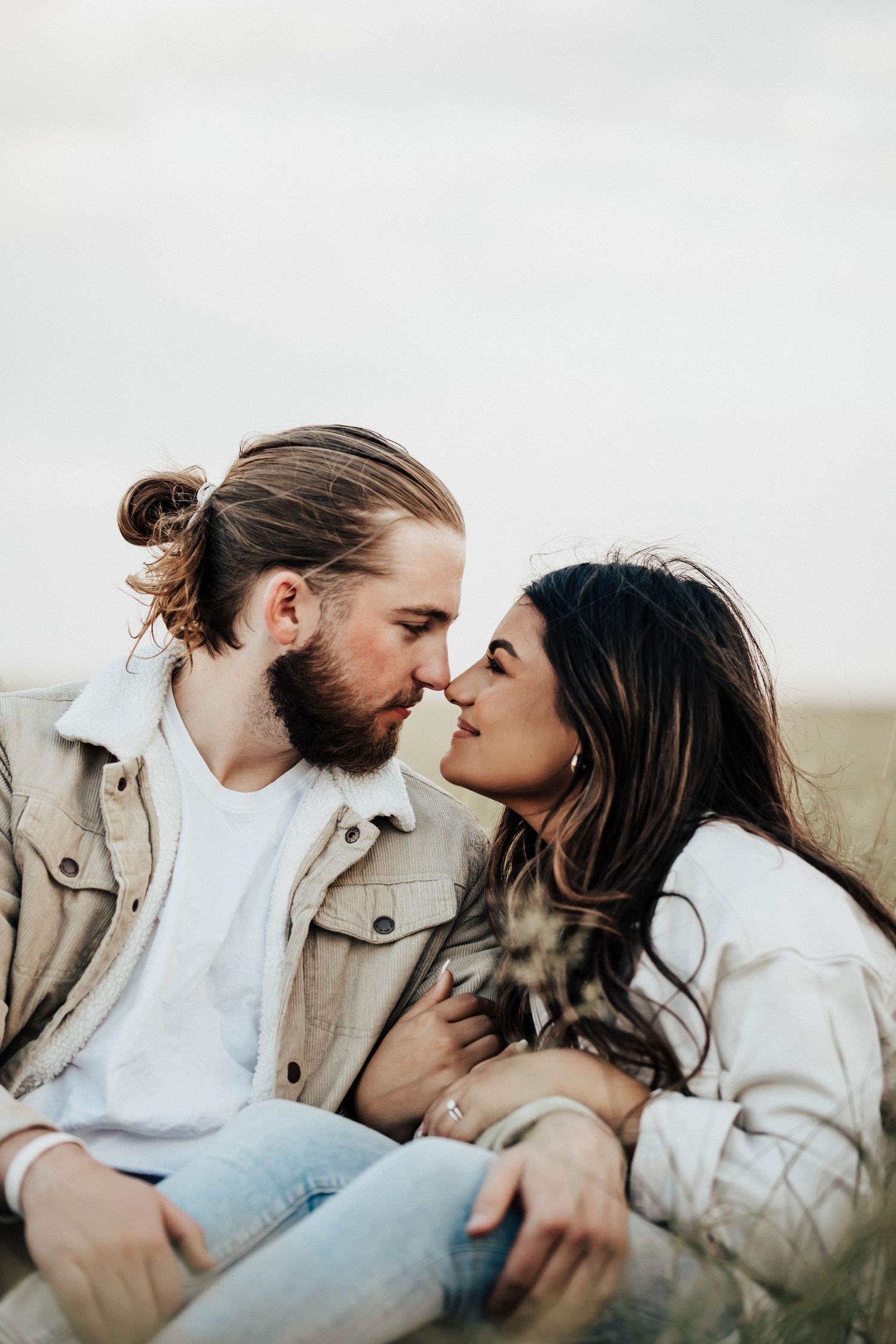 A bearded man and dark-haired woman share a romantic almost-kiss while sitting together in a grassy field.