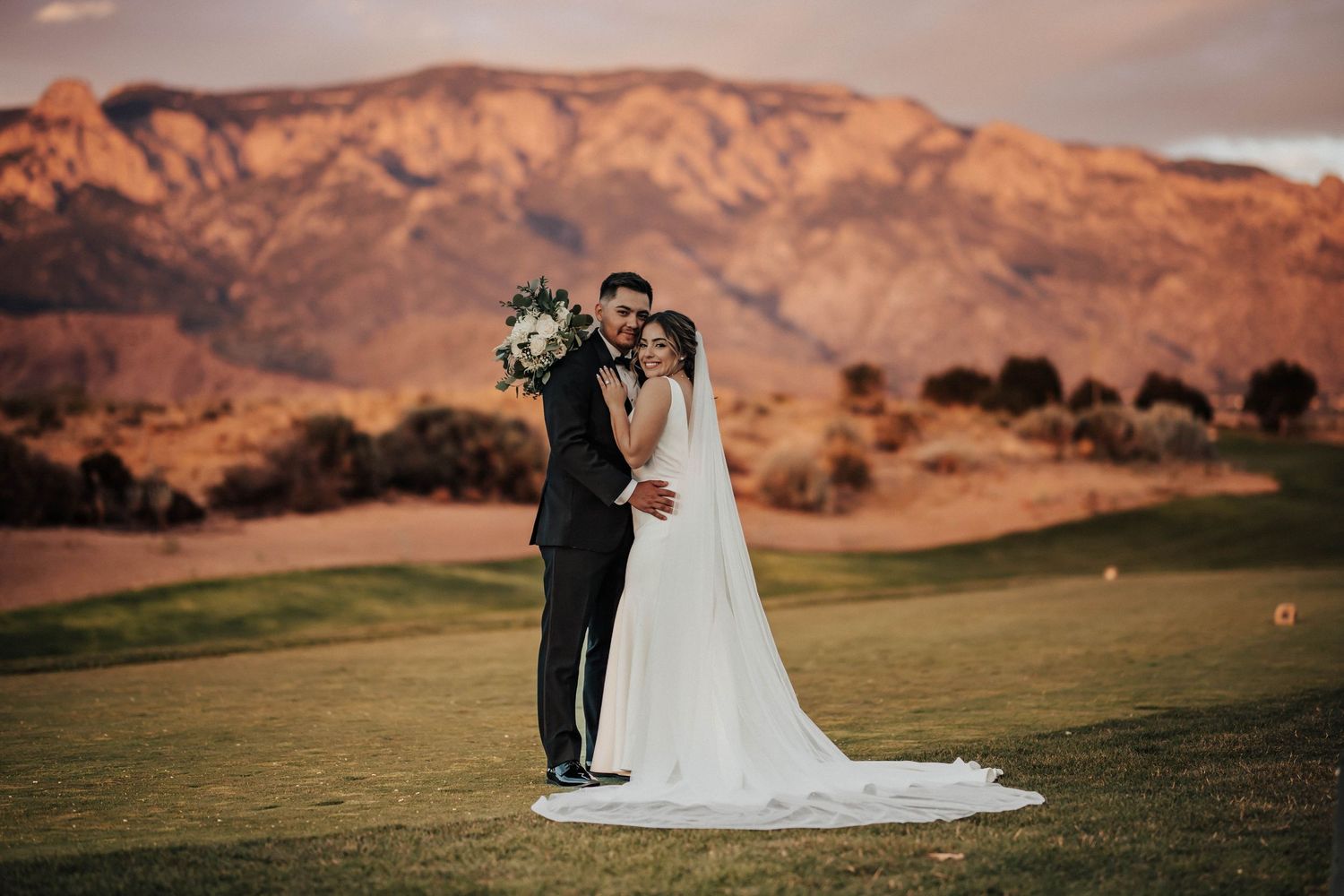 Bride and groom pose on golf course with stunning red mountain backdrop at golden hour sunset wedding.