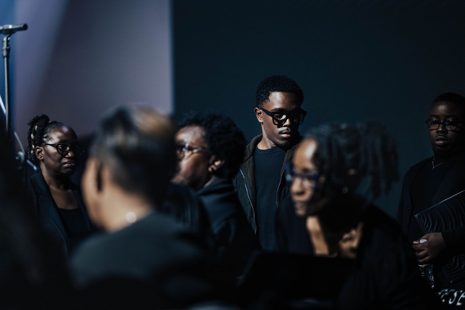 A choir dressed in black performs on stage, with a young man in sunglasses standing prominently in the group.