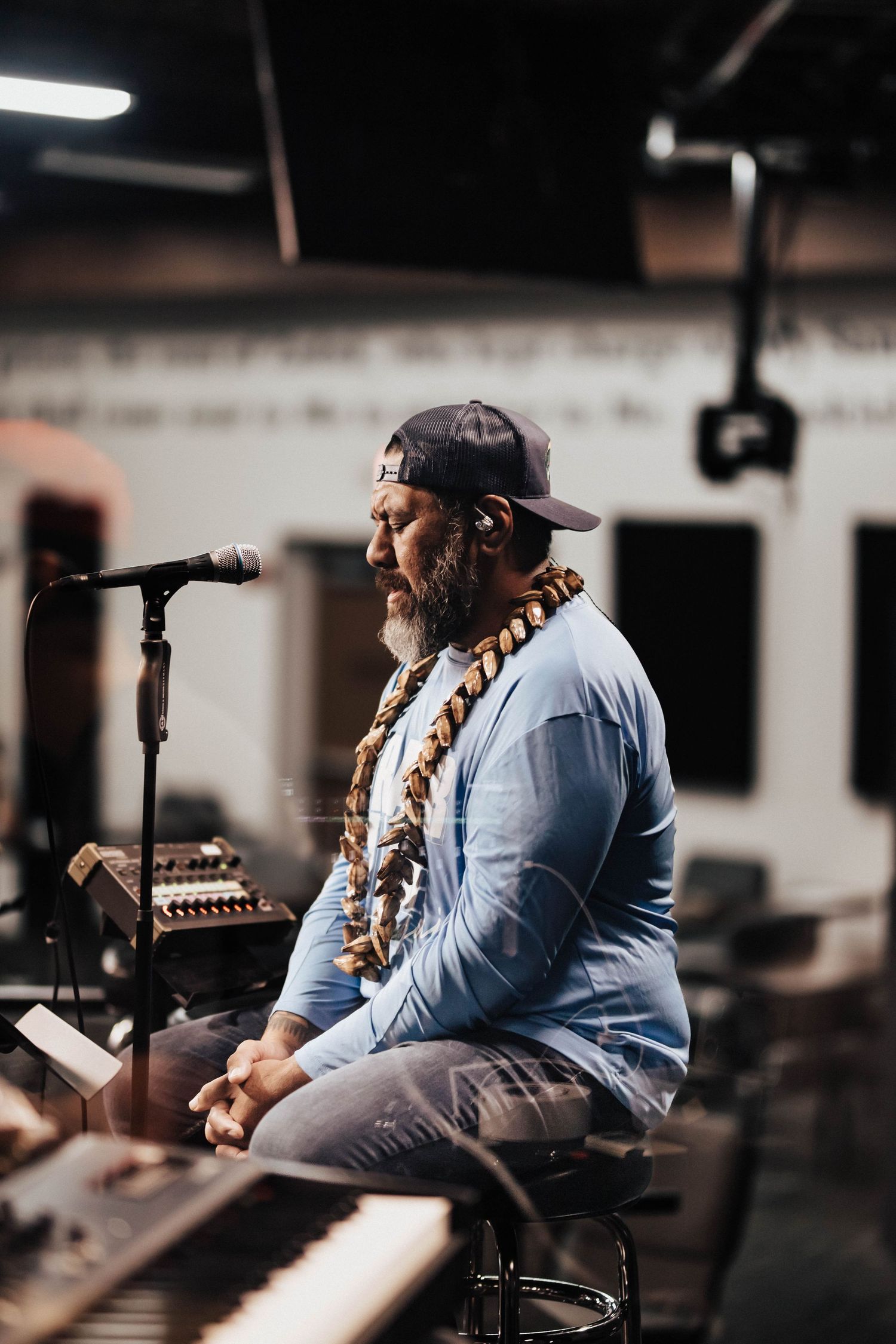 A man wearing a lei and cap sits at a microphone beside a keyboard during a worship session.