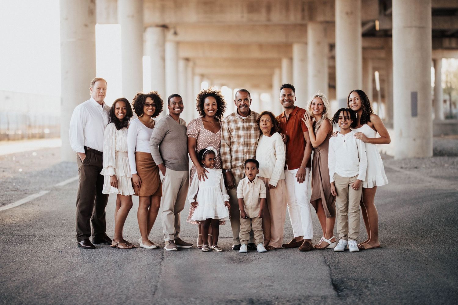 Large multi-generational family posing together under bridge pillars in coordinated neutral outfits.