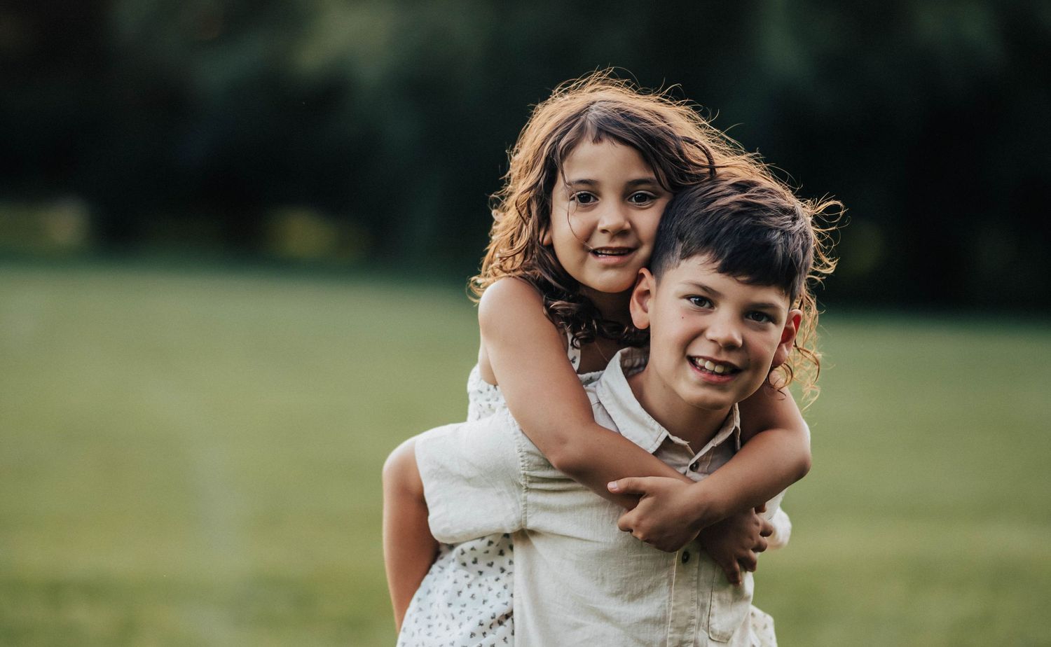 A smiling boy gives a happy girl a piggyback ride in a lush green park during a sunny day.