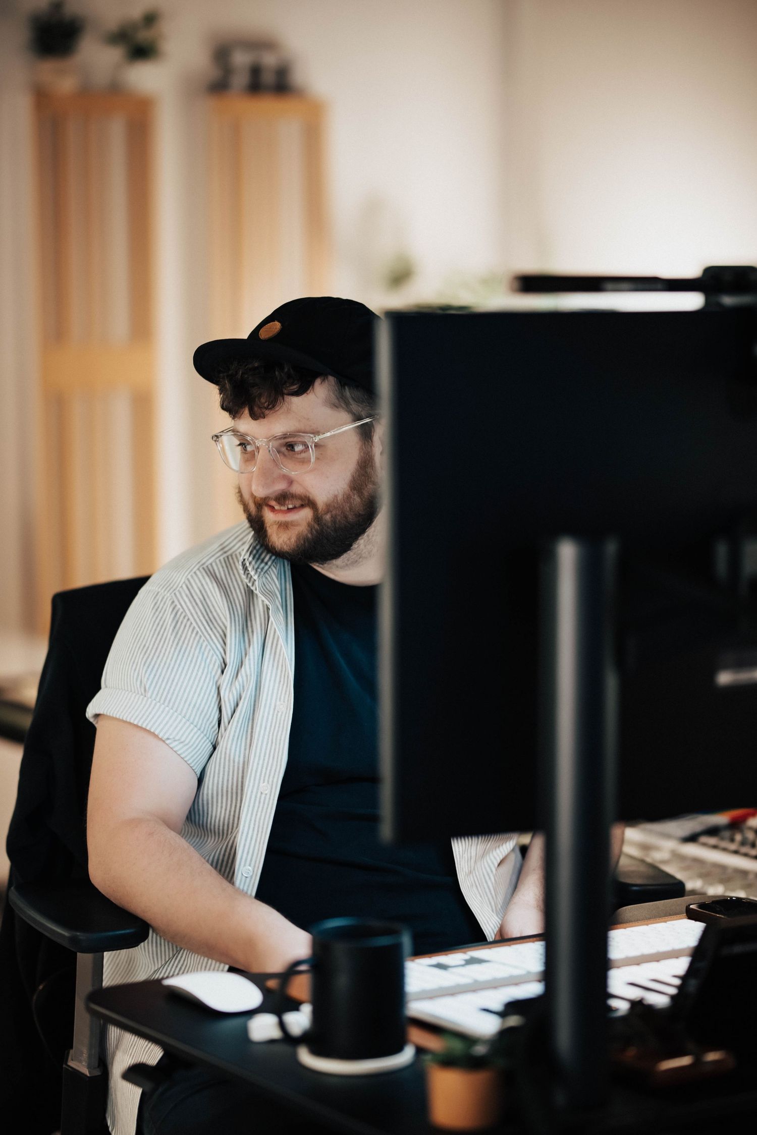 Bearded man wearing glasses and a cap working at a computer desk in a modern office environment.