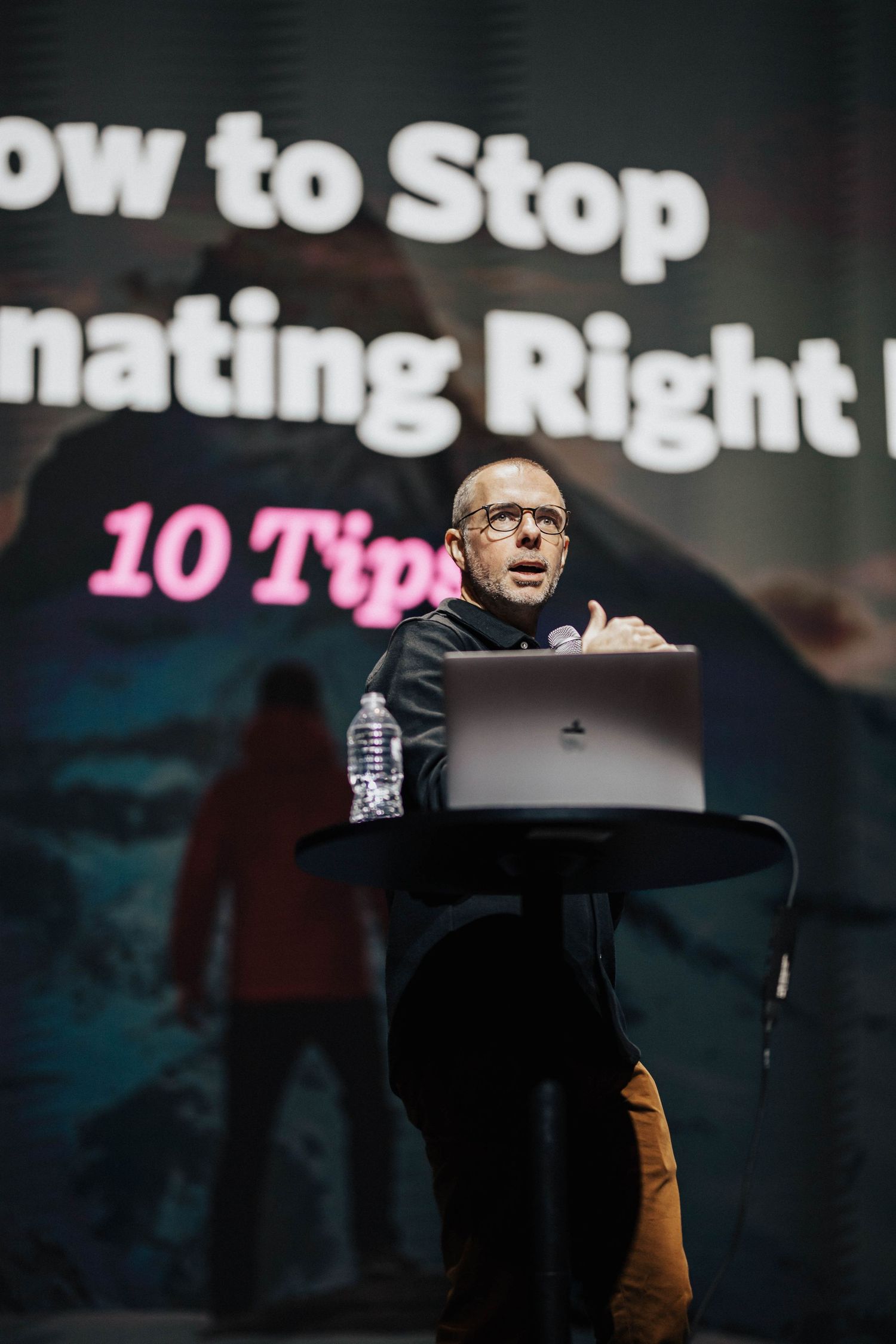 A speaker presents '10 Tips on How to Stop Procrastinating' at a podium with a laptop during a conference.