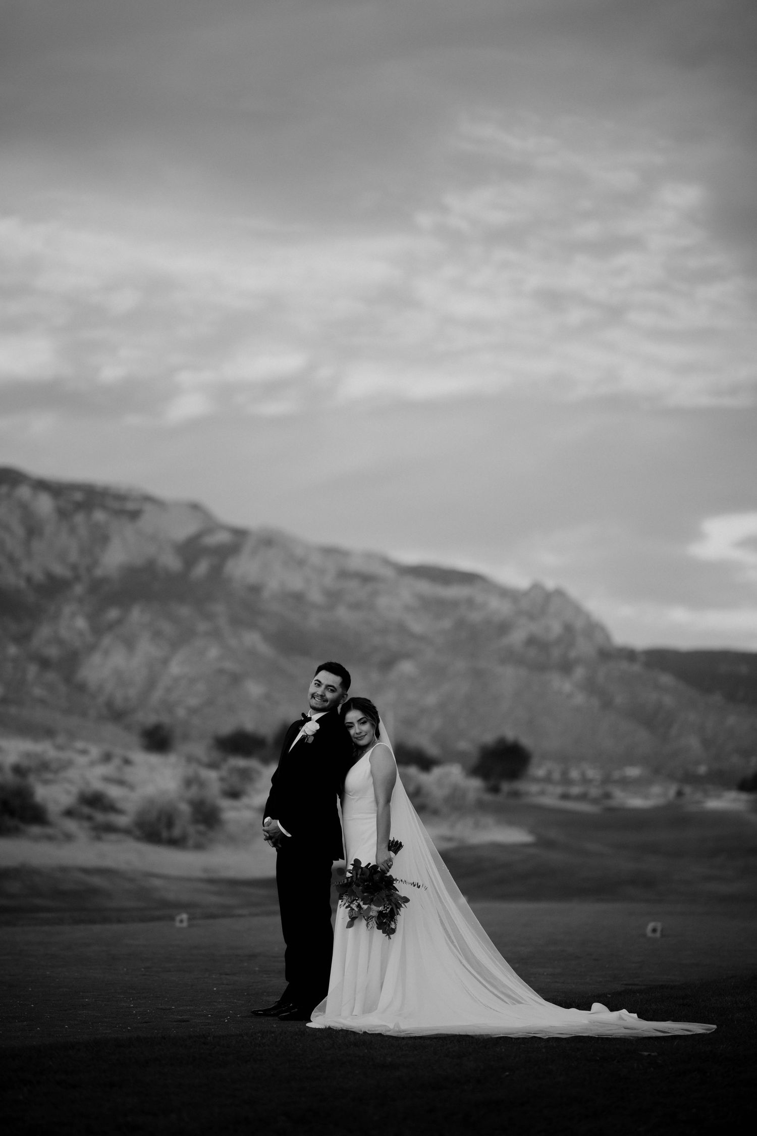 Black and white wedding photo of bride and groom posing outdoors with mountain backdrop and dramatic cloudy sky.